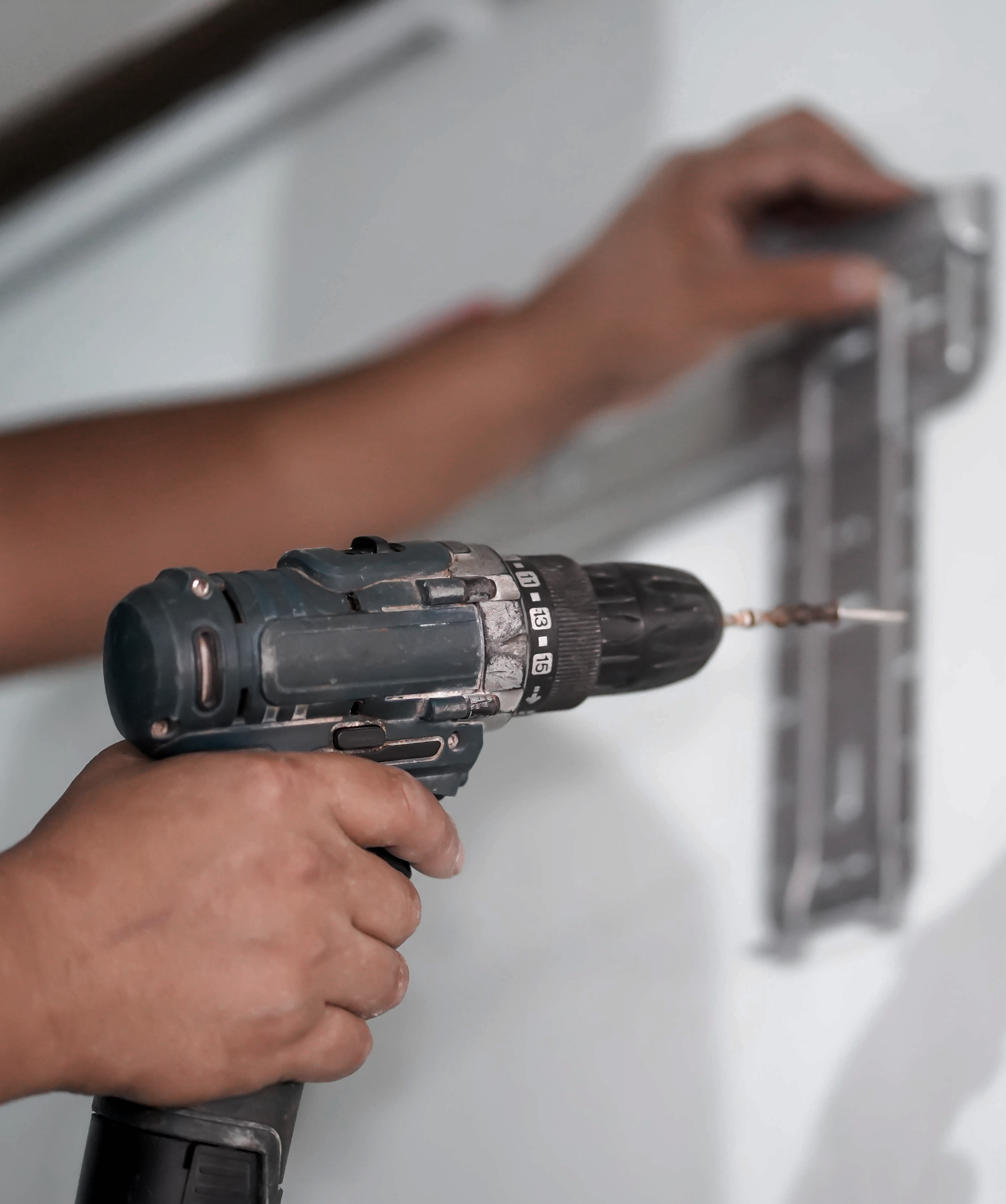 A person holding a cordless power drill with a black and gray body, drilling into a surface, with a gray background.