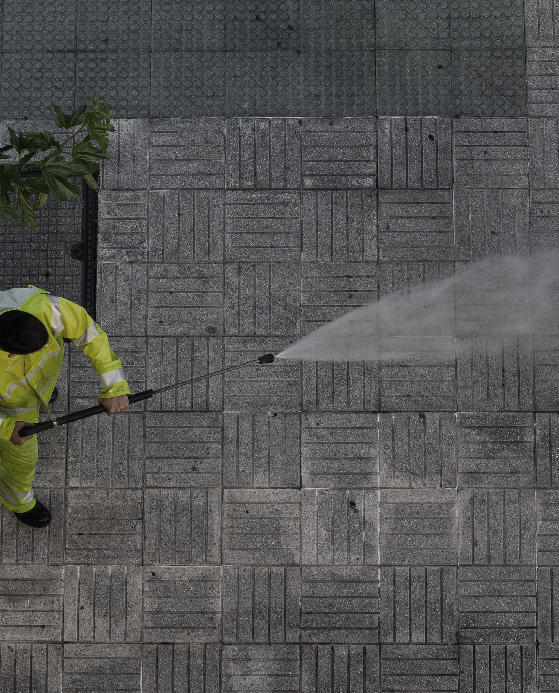 Worker in a yellow safety vest cleaning a sidewalk with a high-pressure water spray.