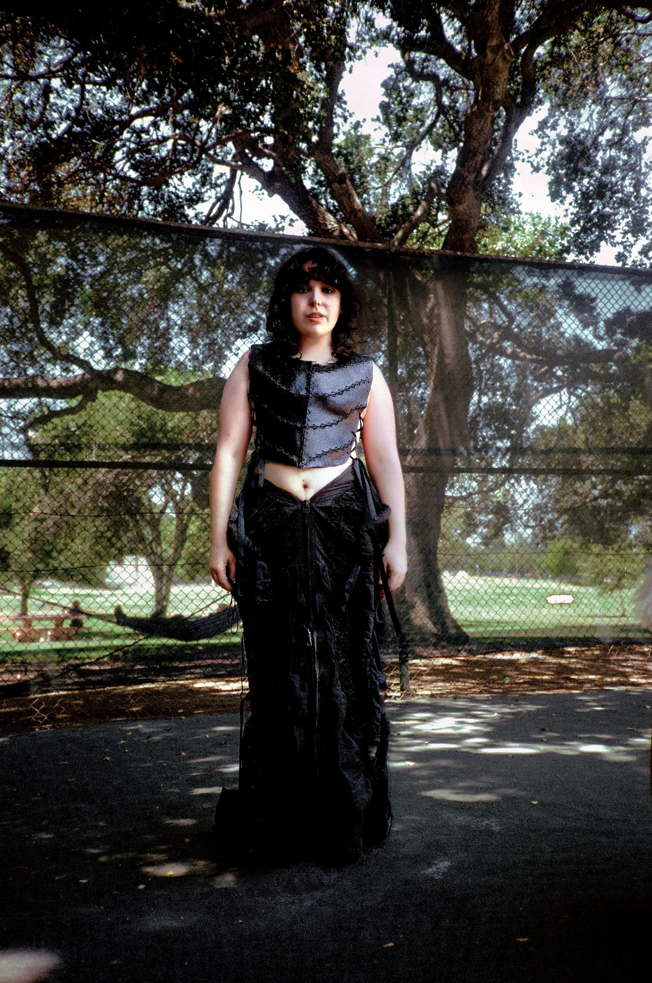 A woman with dark hair, wearing a black shiny top and long black skirt, stands outdoors in front of a chain-link fence and large tree. There are trees and a field in the background, with sunlight filtering through the leaves, casting shadows.