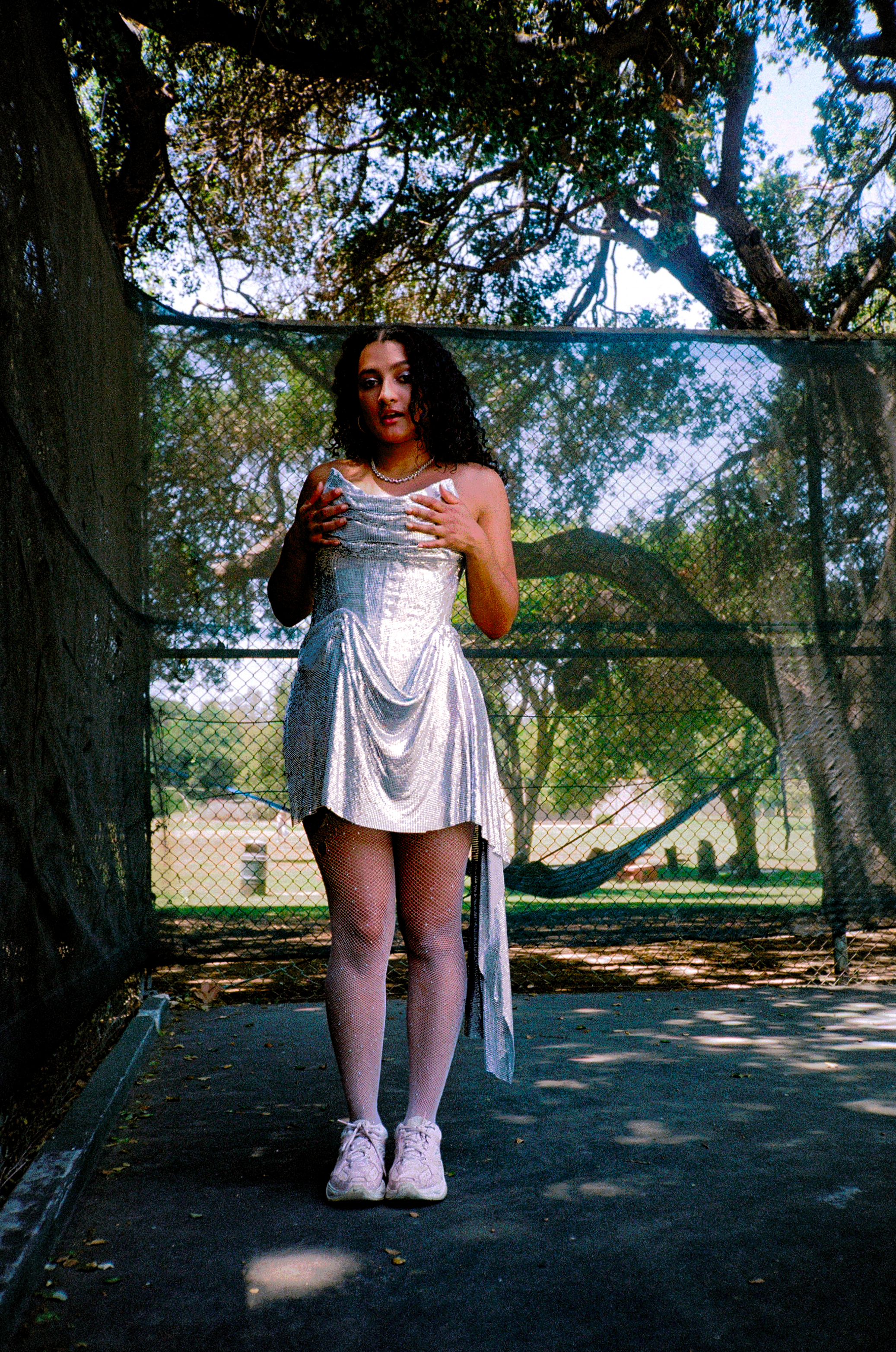 A person with curly dark hair stands in front of a chain-link fence, outdoors, wearing a shiny silver dress, white fishnet stockings, and pink sneakers, with hands on their chest and a neutral expression.