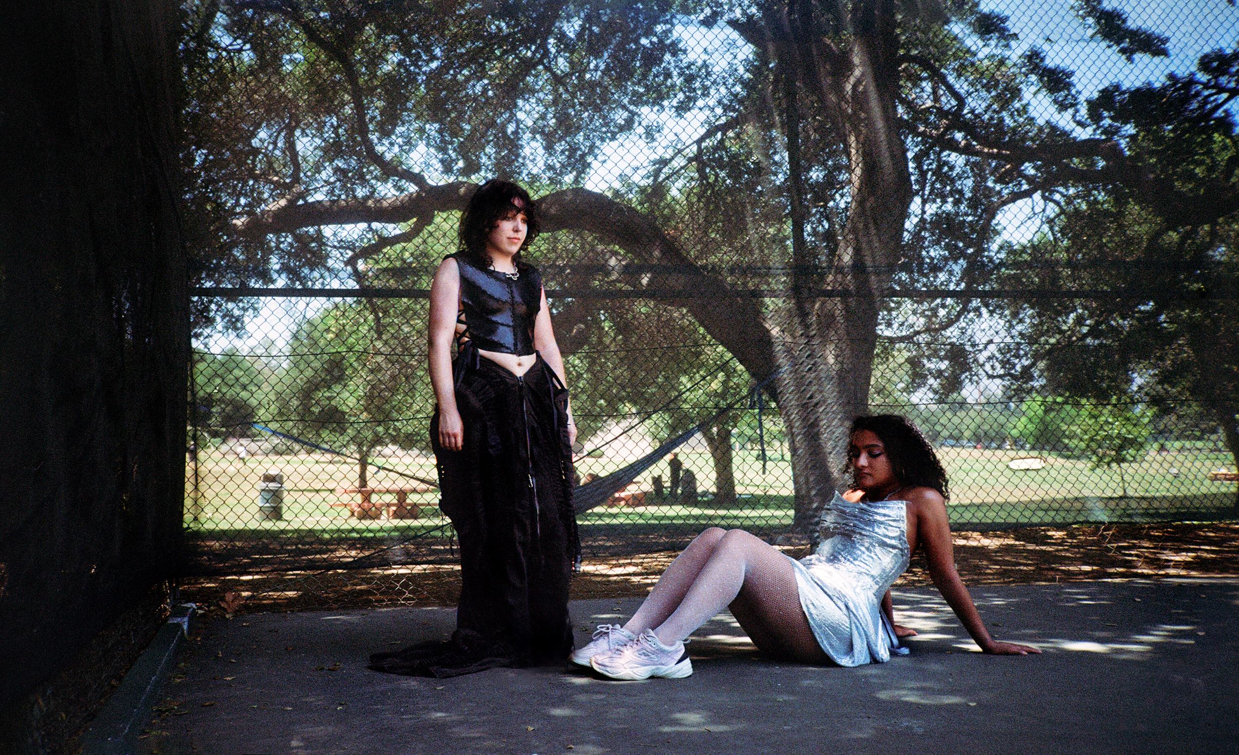 Two young women are in a park enclosed by a chain-link fence, with a large tree and picnic tables in the background. One woman is standing, wearing a black outfit, and the other is sitting on the ground, wearing a shiny silver dress and white sneakers.