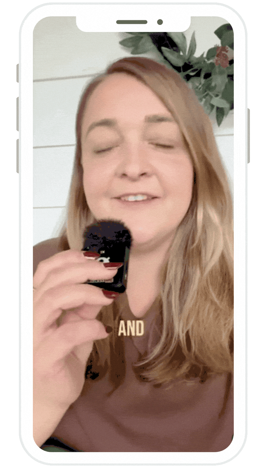 A woman with long, wavy blonde hair holding a microphone with a black fluffy cover, smiling with eyes closed, in front of a white wall with green foliage decor.