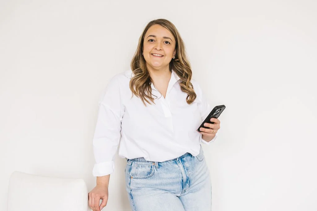 A young woman with light brown hair holding a smartphone, standing against a plain white wall, wearing a white shirt and light blue jeans.