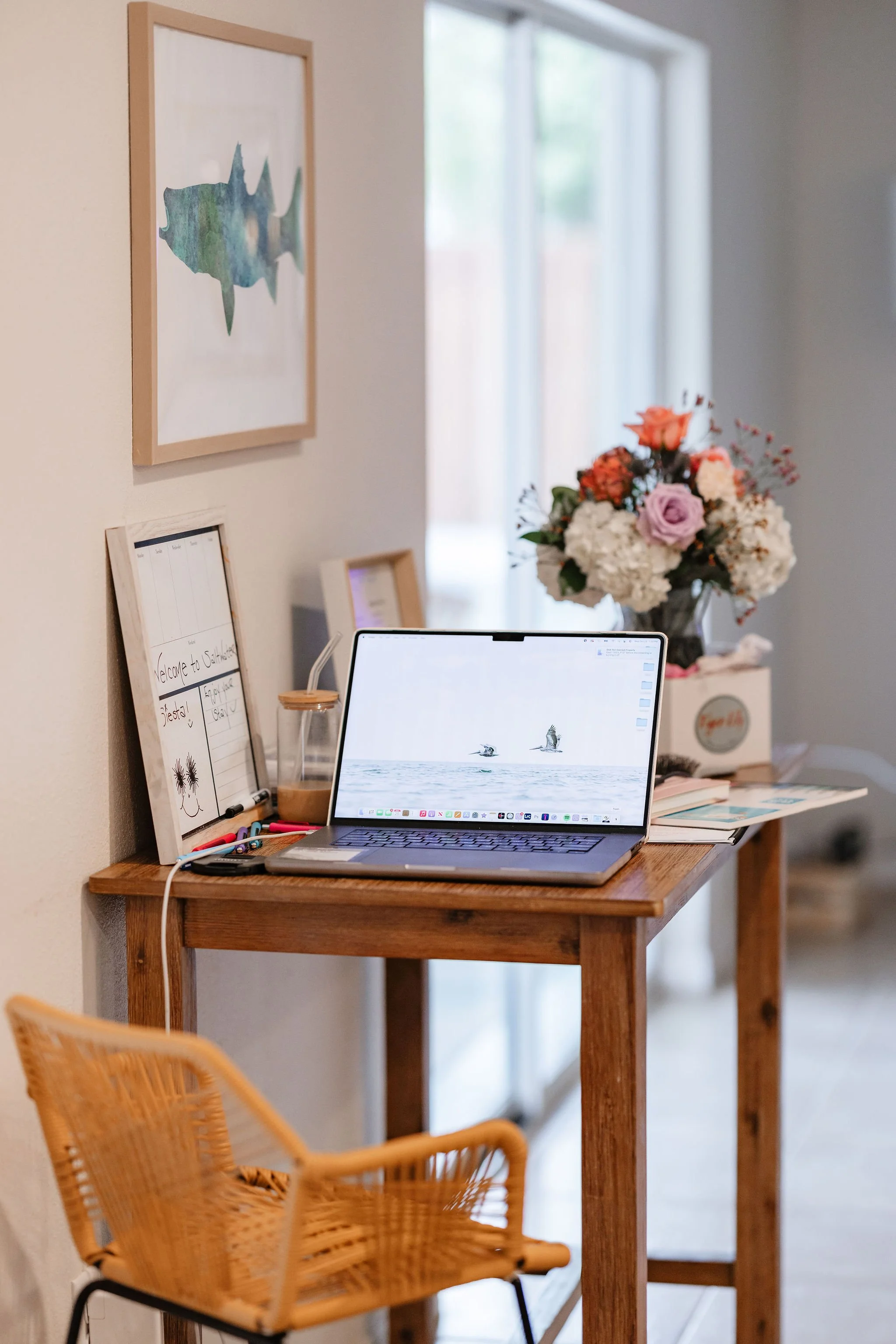 A wooden desk with a laptop displaying a picture of two birds flying over water, a whiteboard with writing, a small framed note, a glass of iced coffee with a straw, and a stack of books. Behind the desk, a vase with a bouquet of mixed flowers and a window with natural light.