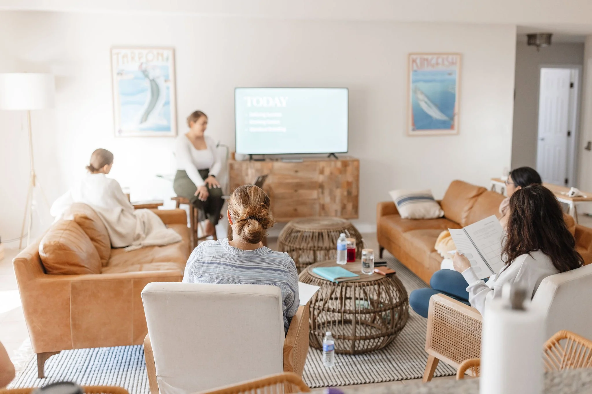 A group of people attending a presentation or meeting in a cozy living room setting. The presenter, a woman, is sitting on a stool near a flat-screen TV displaying a slide titled 'Today'. The audience, including women and a young girl, is seated on sofas, chairs, and an armchair, with some taking notes and water bottles on the tables.
