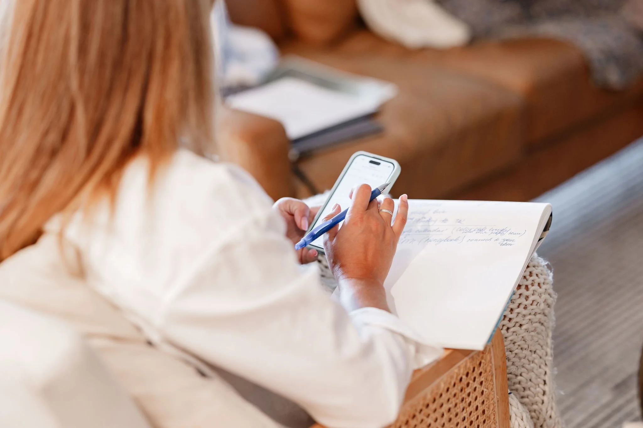 A woman with long, light brown hair is writing in a notebook, holding a pen in her right hand and a smartphone in her left. She is sitting at a wooden table with other notebooks in the background.