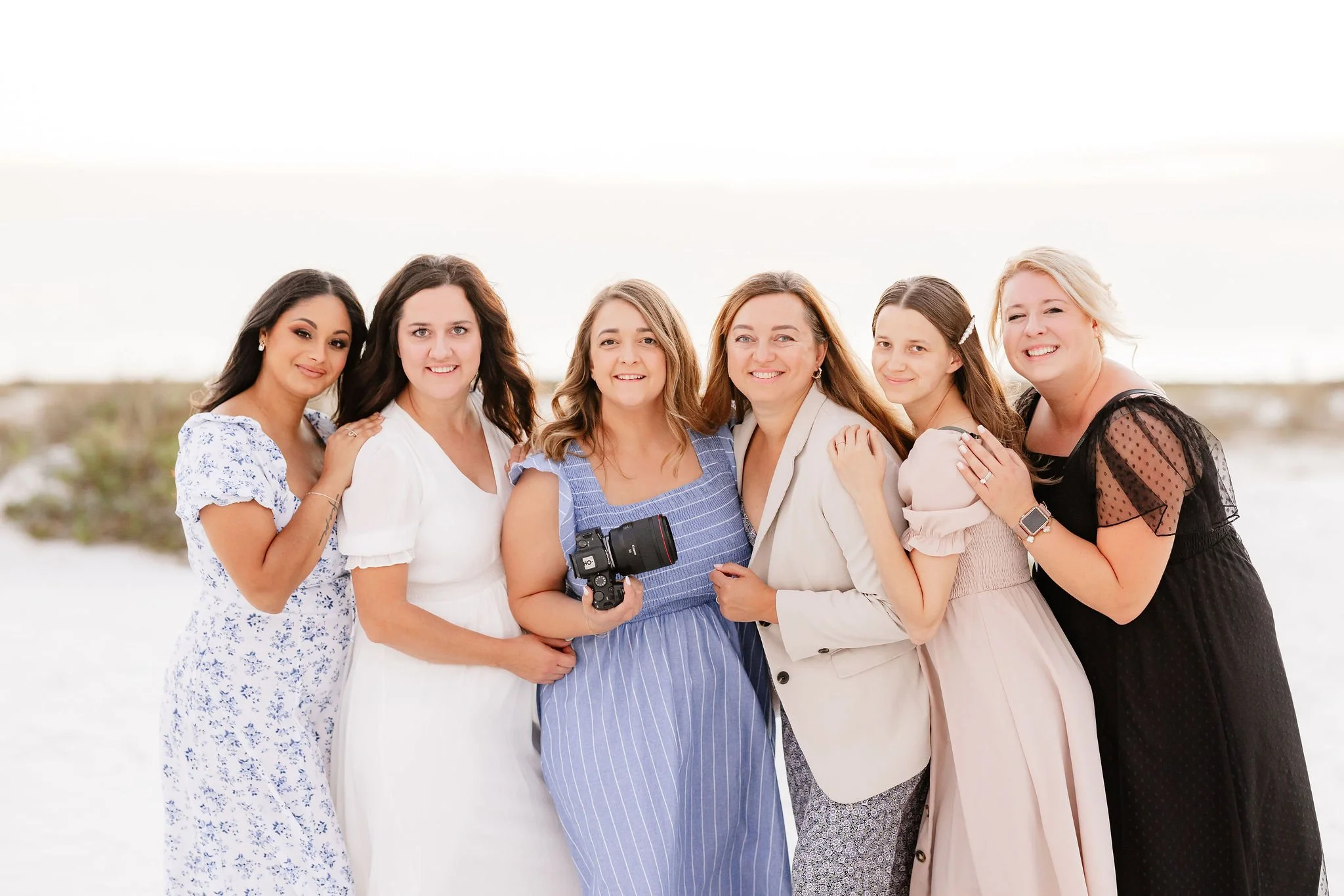 Group of seven women posing together outdoors with a clear, bright sky in the background, dressed in light-colored and patterned dresses, one woman holding a camera.