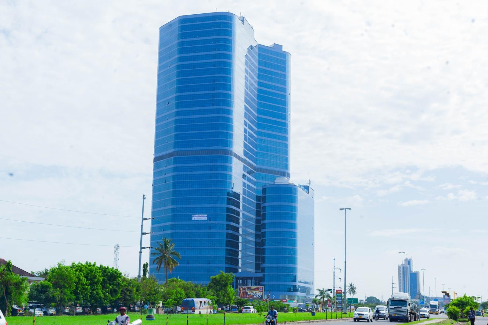 A tall, modern glass skyscraper with blue tinted windows, surrounded by greenery and a busy street with cars, motorcycles, and streetlights.