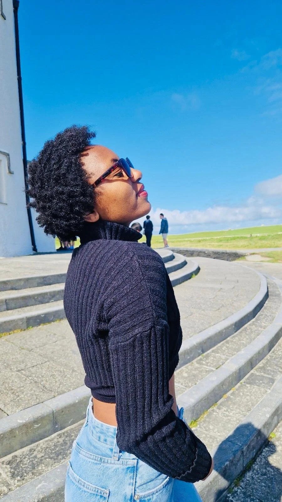 Young woman with curly hair, wearing sunglasses, a black sweater, and light blue jeans, standing outdoors on concrete steps, looking up at the sky on a sunny day.