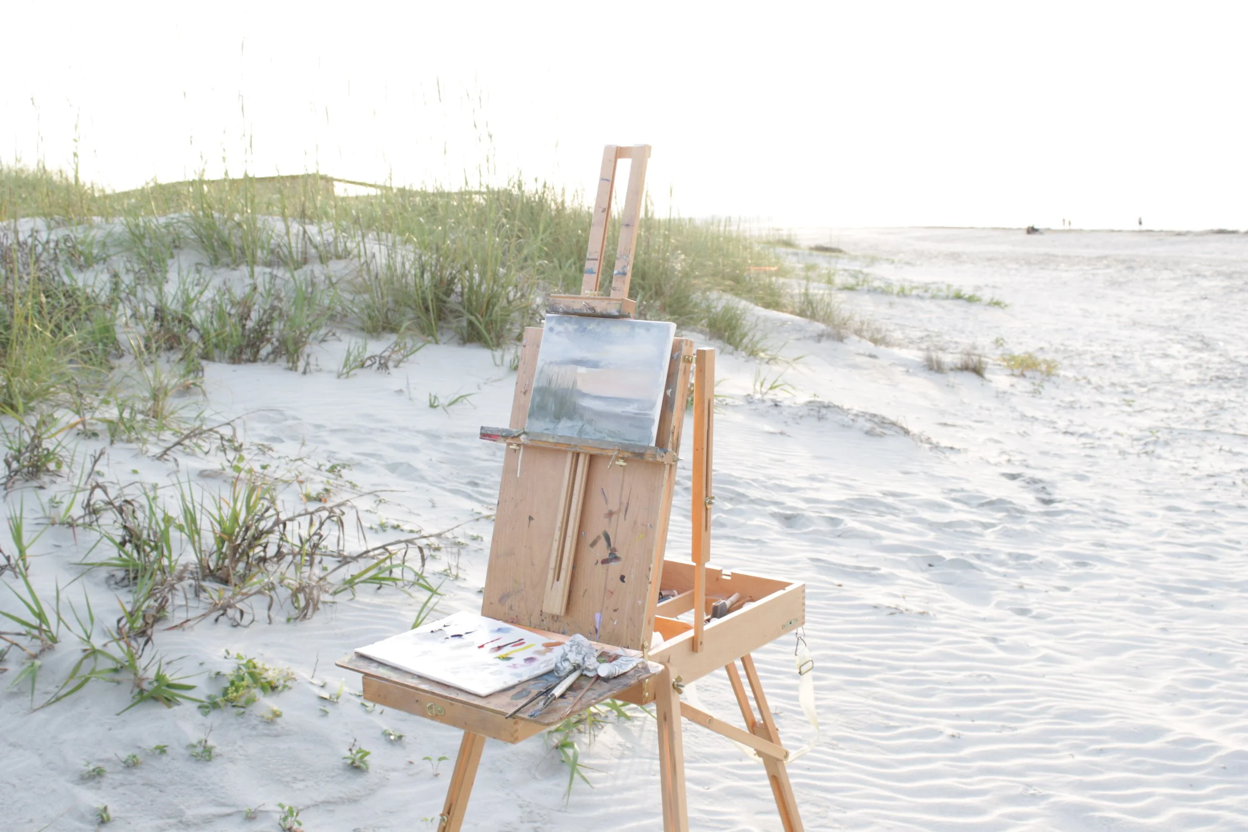 An outdoor paint easel with a partially painted landscape scene sits on a sandy beach with grass in the background.