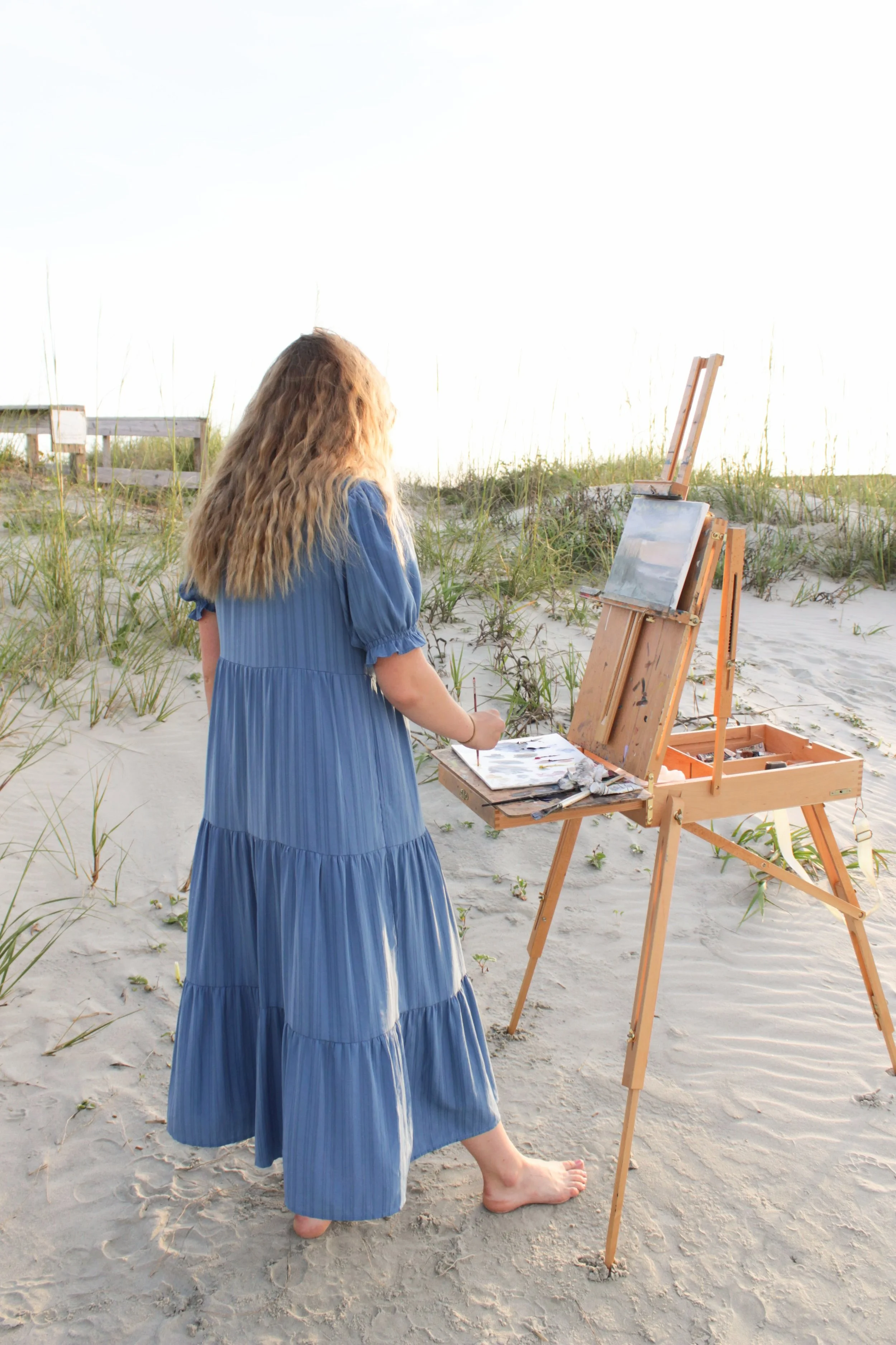 A woman with long, wavy blonde hair wearing a blue maxi dress is standing barefoot on the sand in a beach setting. She is painting on a canvas set up on an easel, with another upright easel nearby. There is sparse grass and a wooden bench in the back
