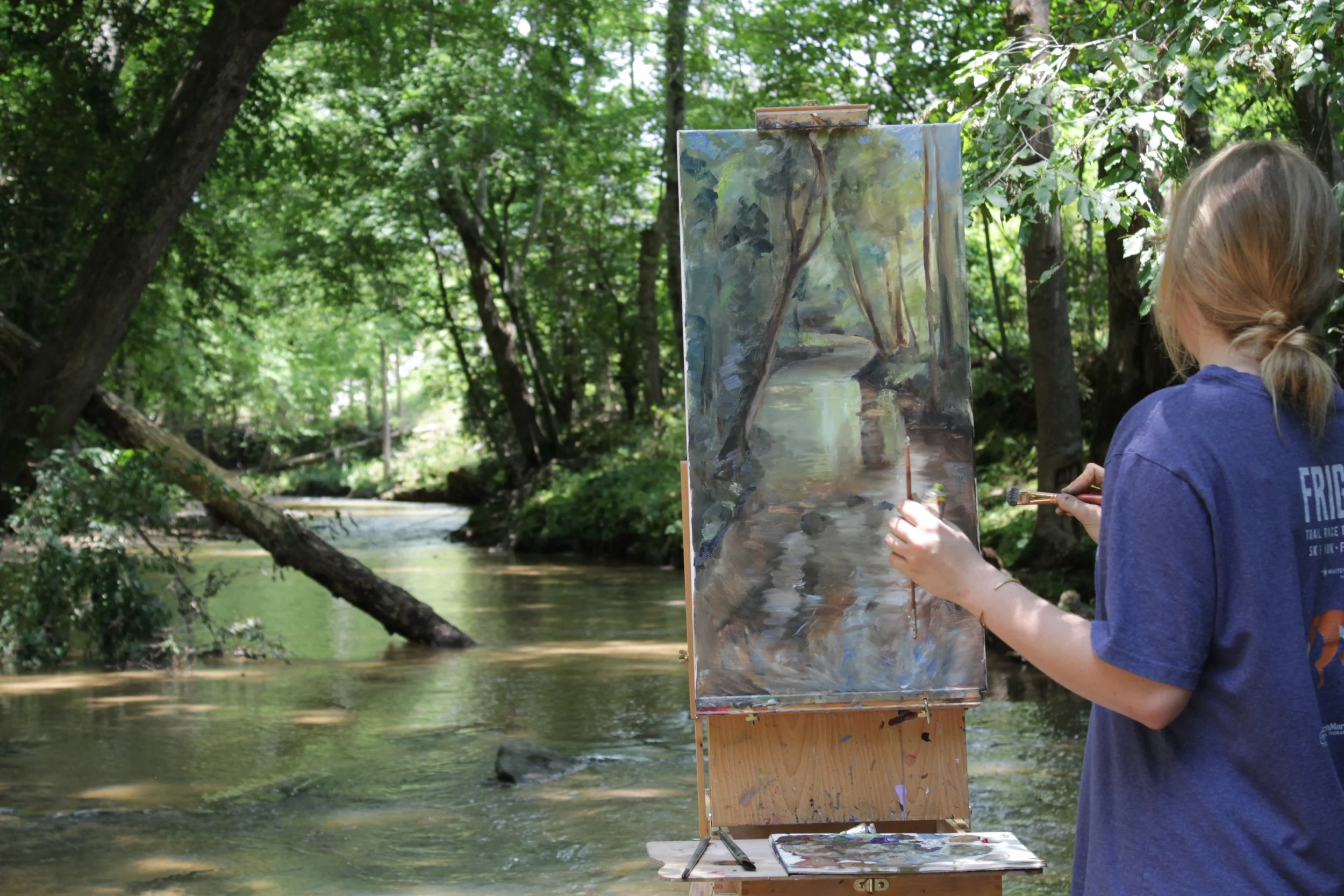 A person painting a landscape of trees and a river on an easel outdoors by a creek in a wooded area.