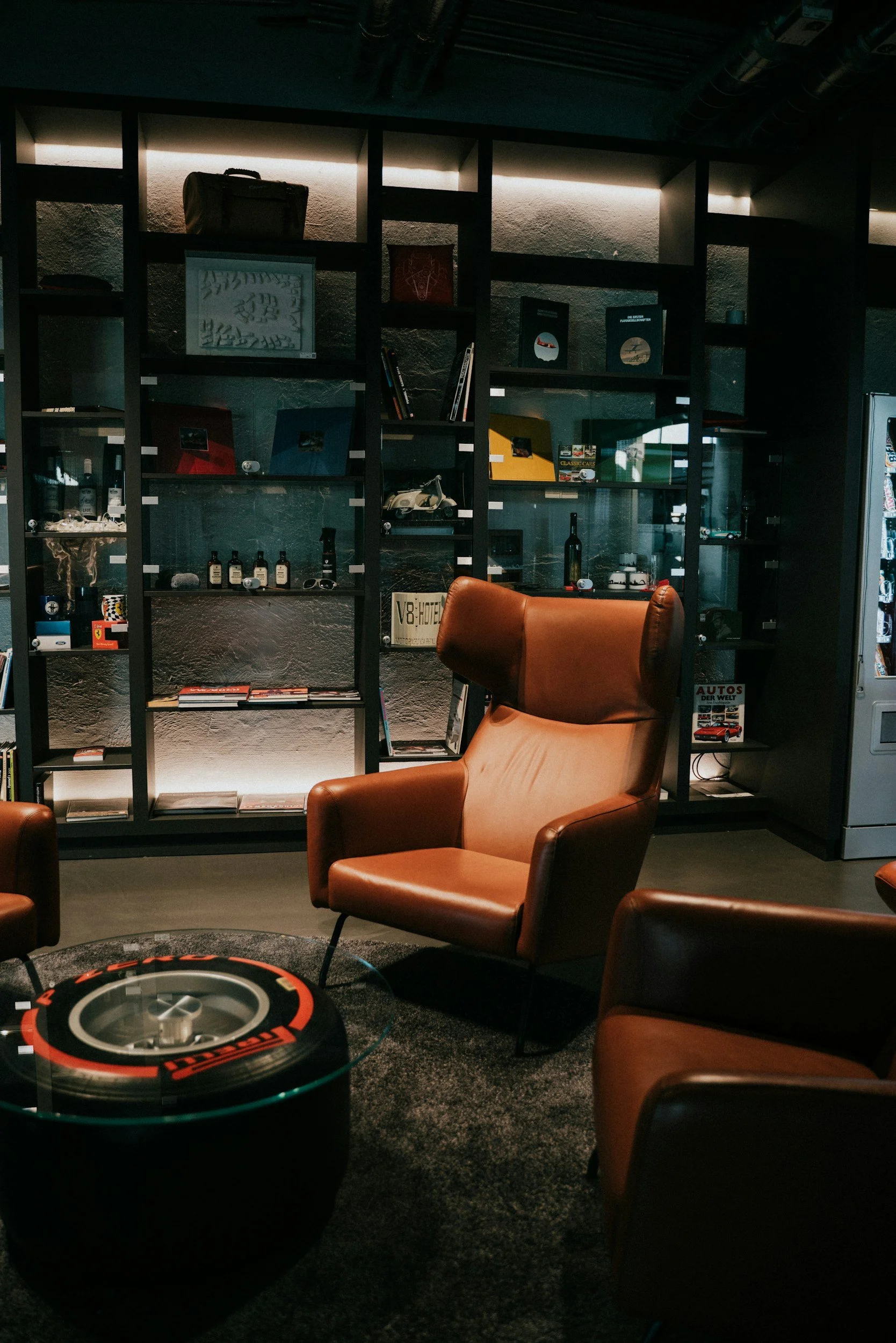 A cozy lounge area with several brown leather armchairs arranged around a small coffee table. Behind the chairs, there is a black shelving unit with various decorative items, books, and framed pictures. The room has dim lighting, creating a warm ambiance.