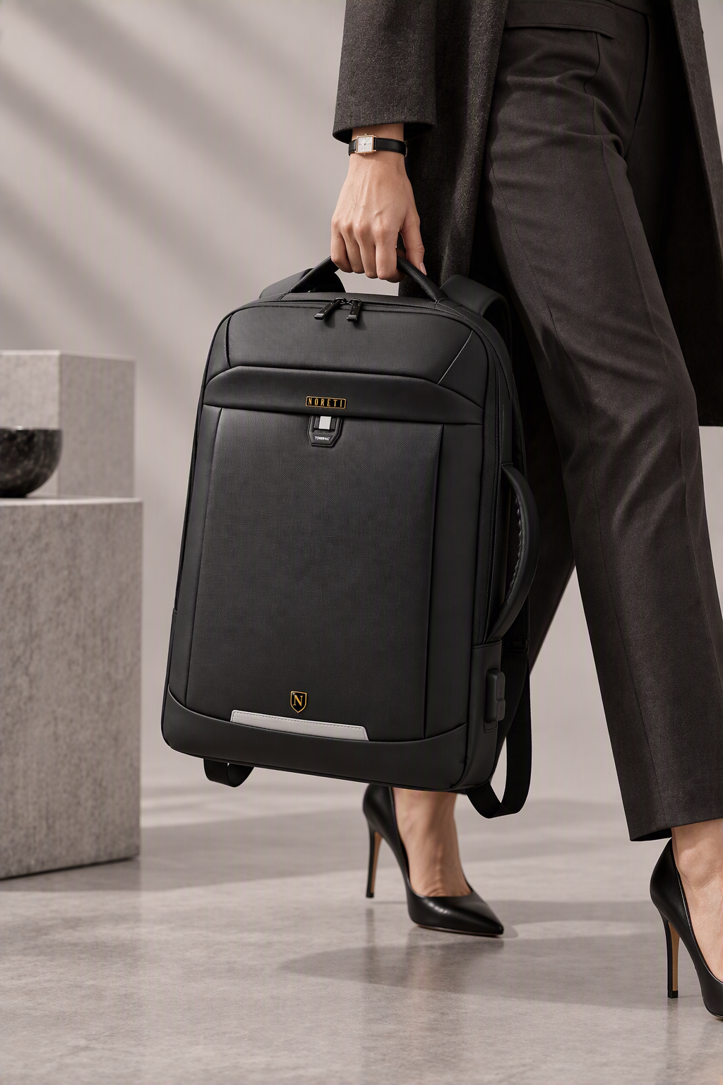 A person in black high heels and black dress pants holds a black rolling briefcase, dressed in a dark blazer in an indoor setting.