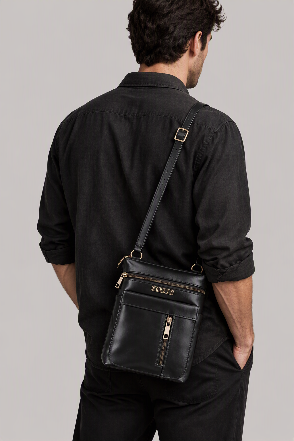 A man in a black shirt carrying a black leather crossbody bag with gold accents, facing away from the camera.