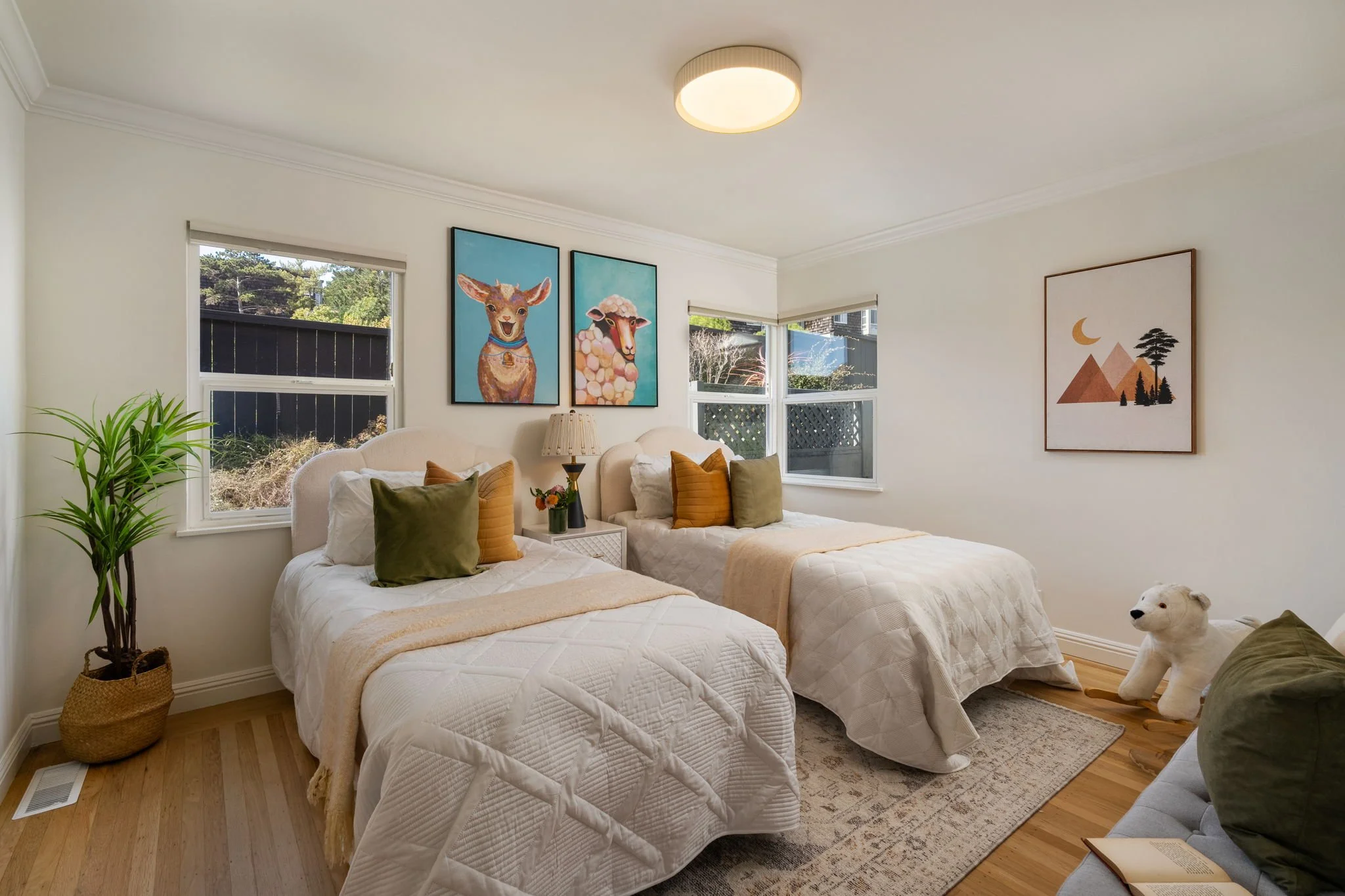 Bedroom with two twin beds, decorated with pillows, a bedside table with a lamp, a potted plant, and artwork on the walls, including images of a deer and a sheep, with windows showing an outdoor scene.