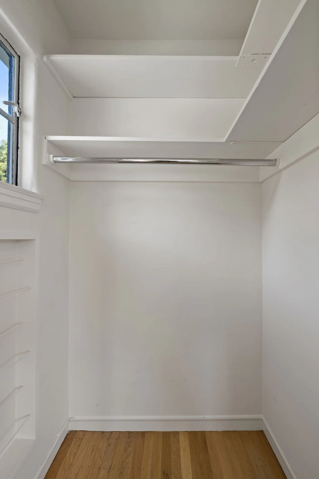 Empty white closet with a metal rod for hanging clothes, a small window on the left, and a built-in white storage unit on the right, with wooden flooring.