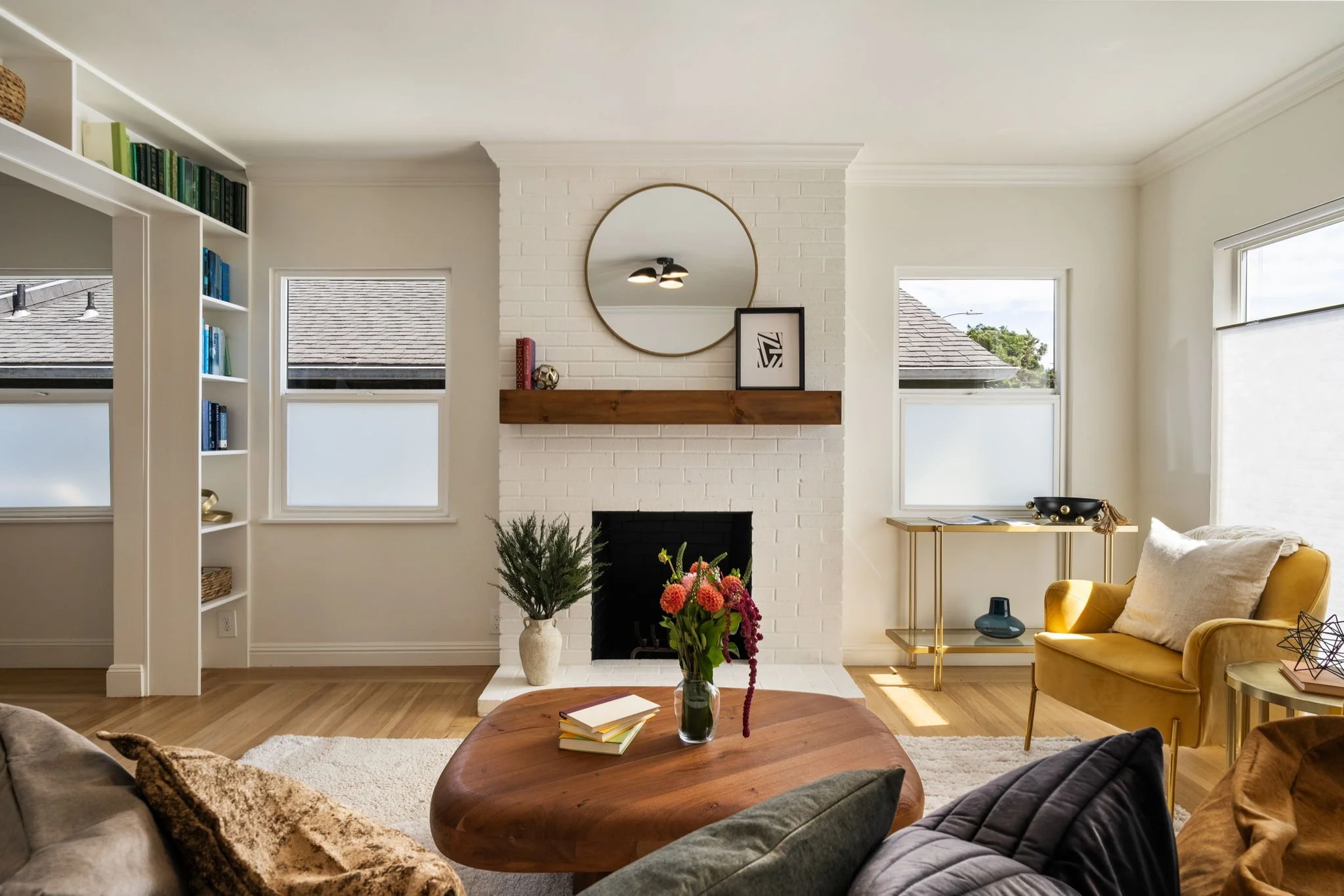 Living room with a white brick fireplace, a round mirror above the mantel, and large windows with blinds. There is a yellow chair, a gold and glass side table, a coffee table with books and a vase of flowers, and built-in shelves with books and decor