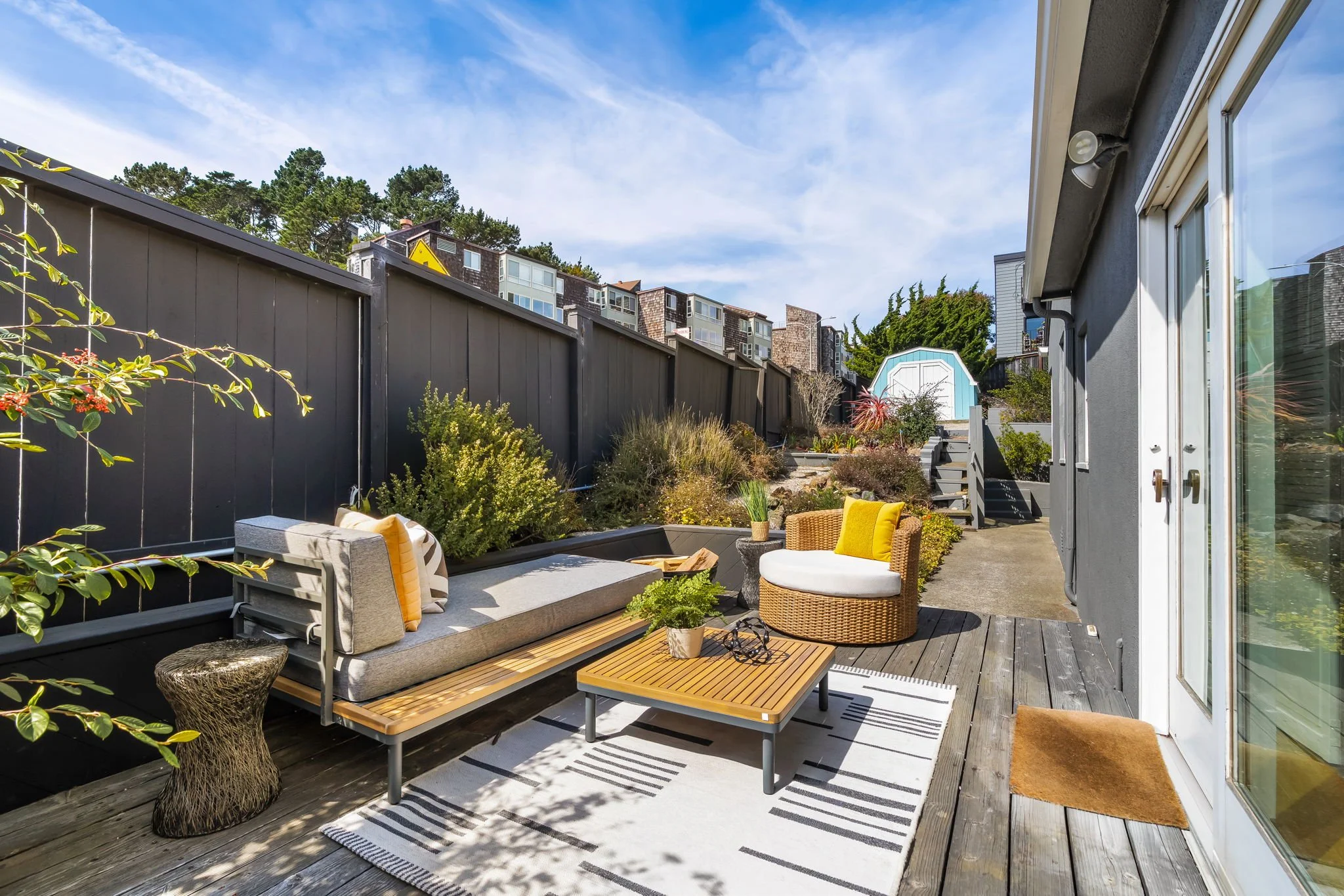 Outdoor patio with seating area including a sectional sofa with cushions, a round wicker chair, a wooden coffee table, and a small side table with plants. There is a striped outdoor rug on a wooden deck, plants along the fence, and a view of neighbor