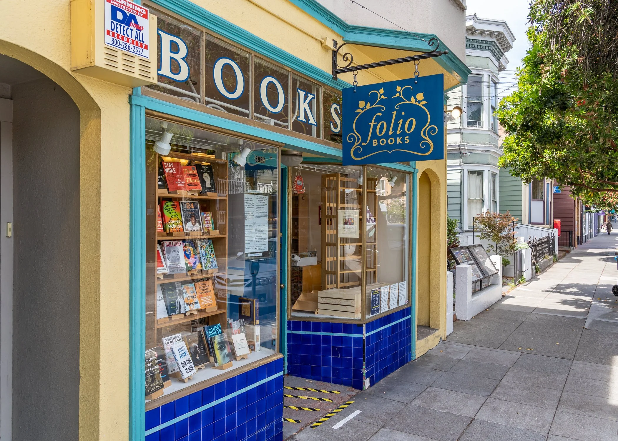 Exterior view of Folio Books, a bookstore with large window display of books and a hanging sign with the store's name. The building has yellow walls and blue trim, and there are trees and other houses on the street visible in the background.