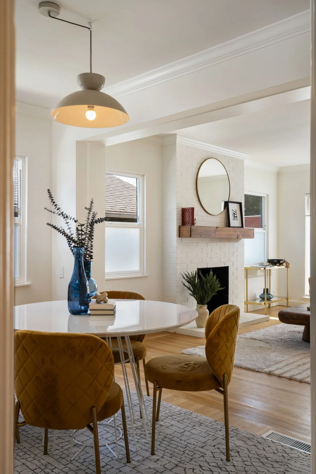 Inside a living room with a round dining table, mustard-colored chairs, a blue vase with branches, a fireplace with a wooden mantel, a round mirror above it, and a beige rug.