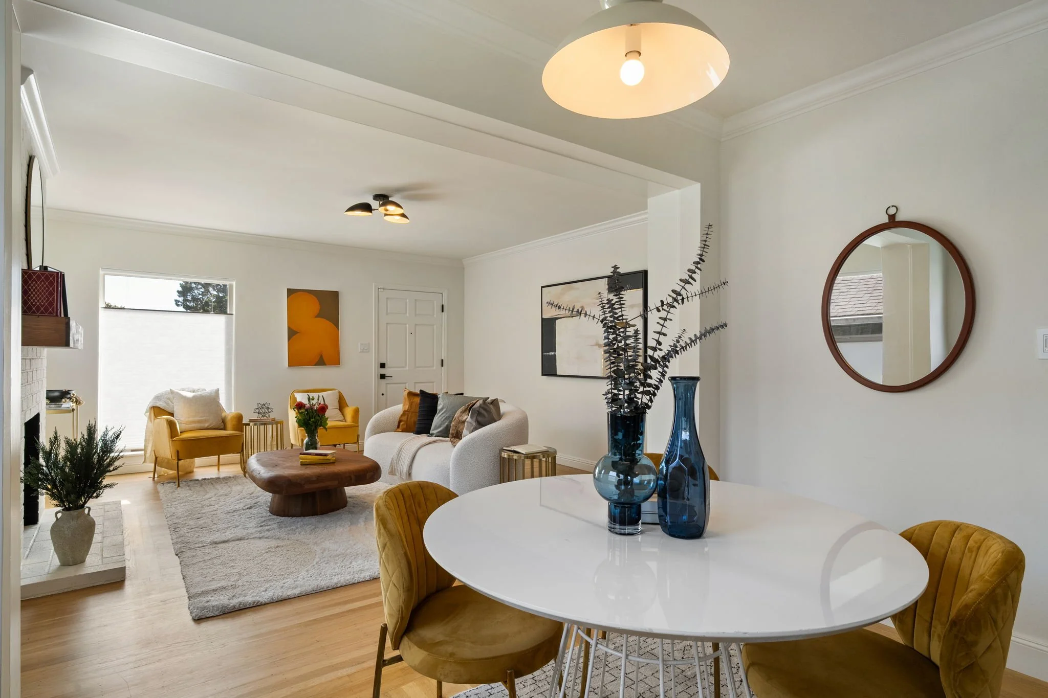 A bright living room with white walls, a round dining table, yellow velvet chairs, a white sofa, a plush gray rug, and wall art, with blue vases and a mirror on the wall.