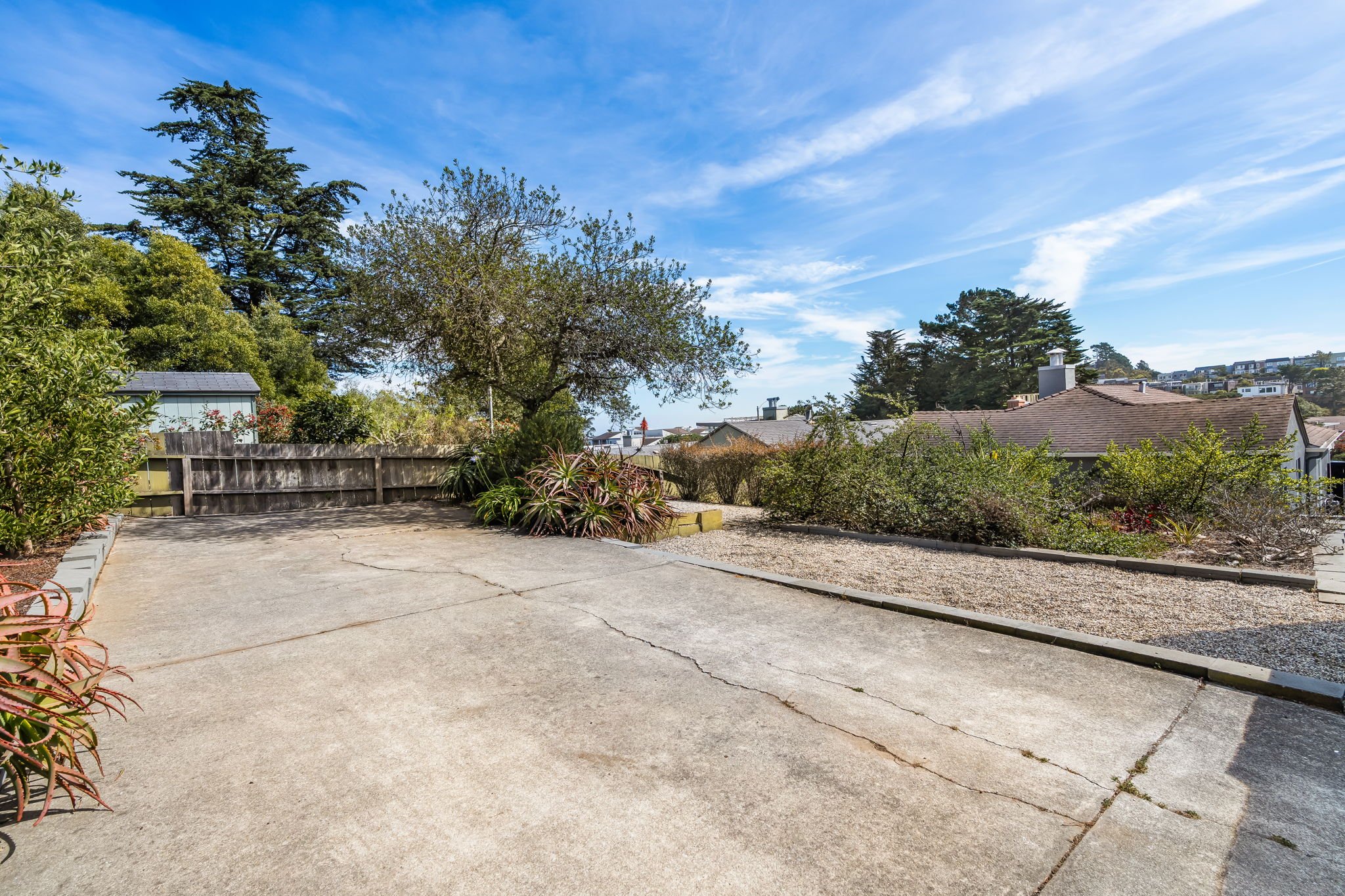 A backyard with a concrete patio, a wooden fence, various bushes and trees, and neighboring houses under a blue sky with wispy clouds.