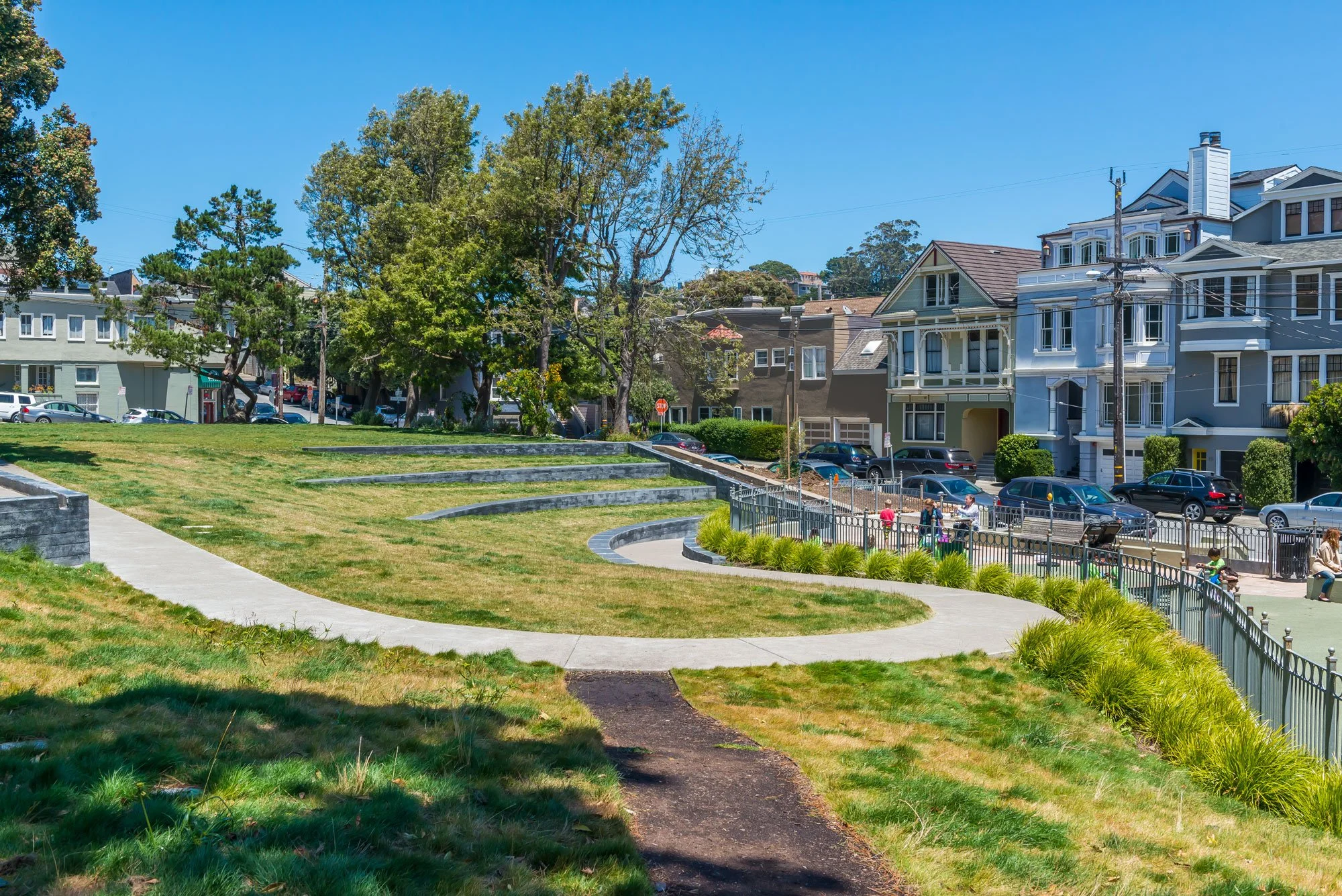 A park with a dirt walking path winding through green grass and a border of yellow-green plants. In the background, there are tall trees, a playground area with children, and residential buildings against a bright blue sky.