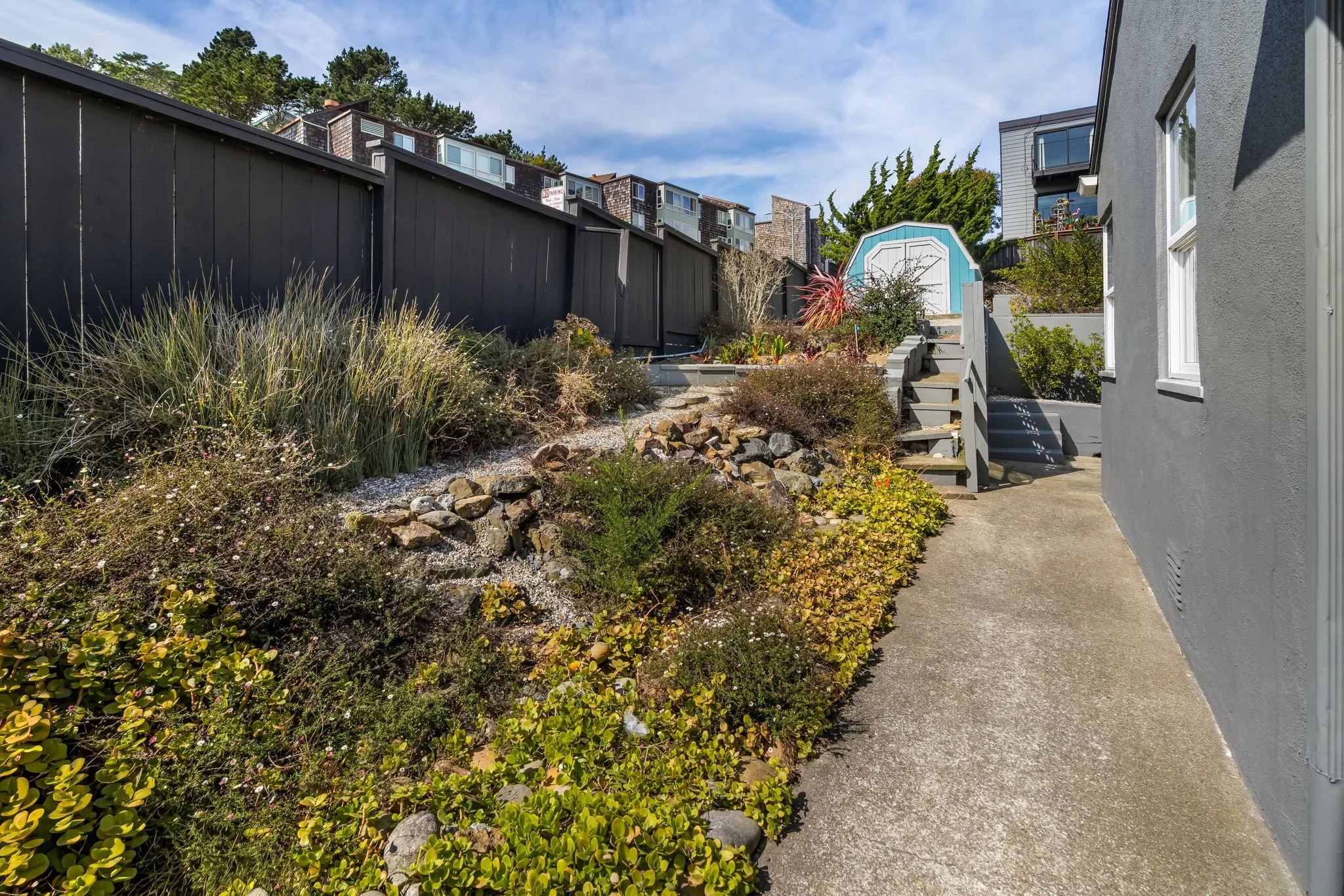 Residential backyard with a sidewalk, garden beds, and a blue storage shed, surrounded by a gray house wall and a black fence, with neighboring apartment buildings in the background.