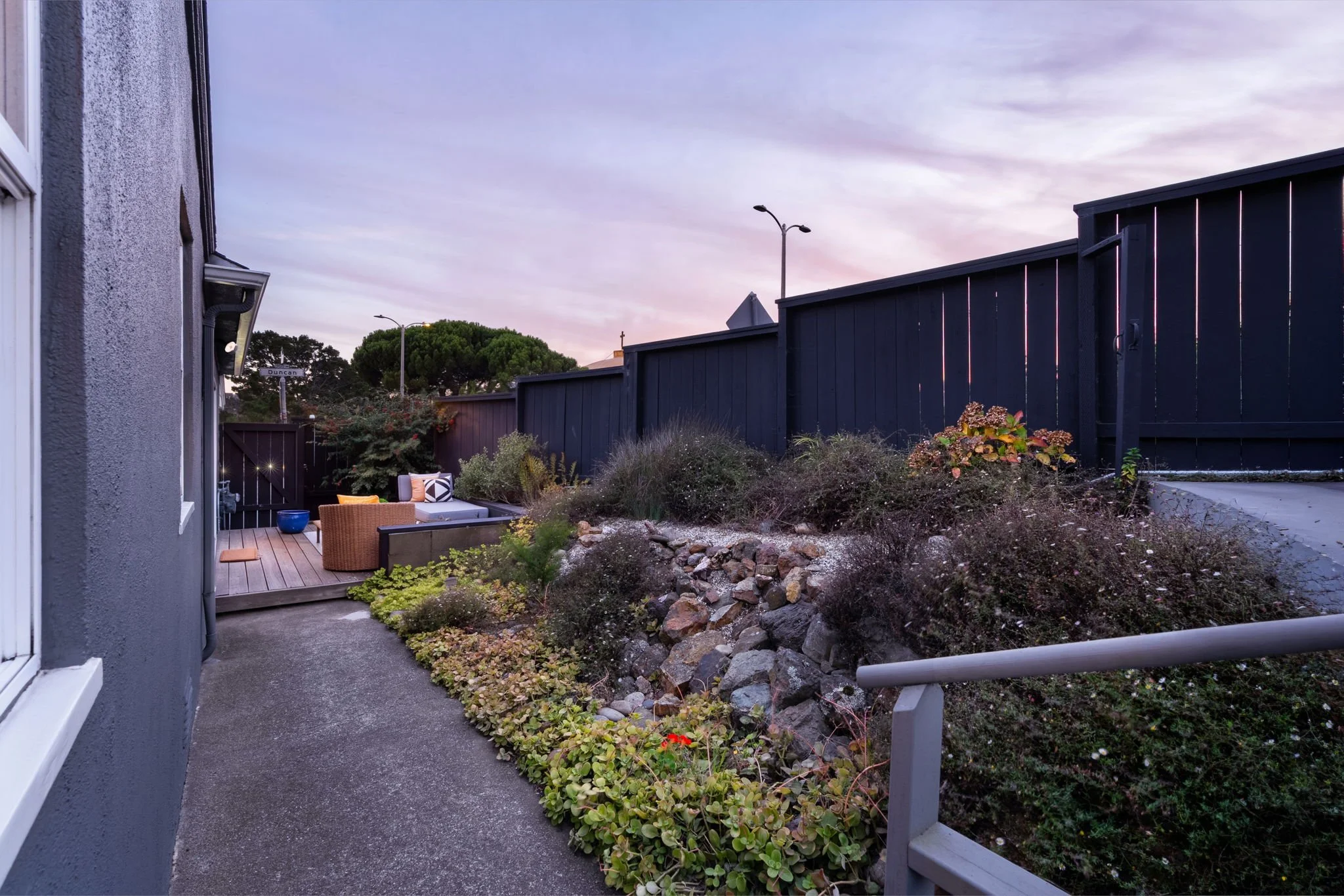 Residential backyard with a concrete pathway, a wooden deck seating area with cushions, surrounded by bushes and rocks, a black wooden privacy fence, a gray house wall with a window, and trees under a partly cloudy evening sky.