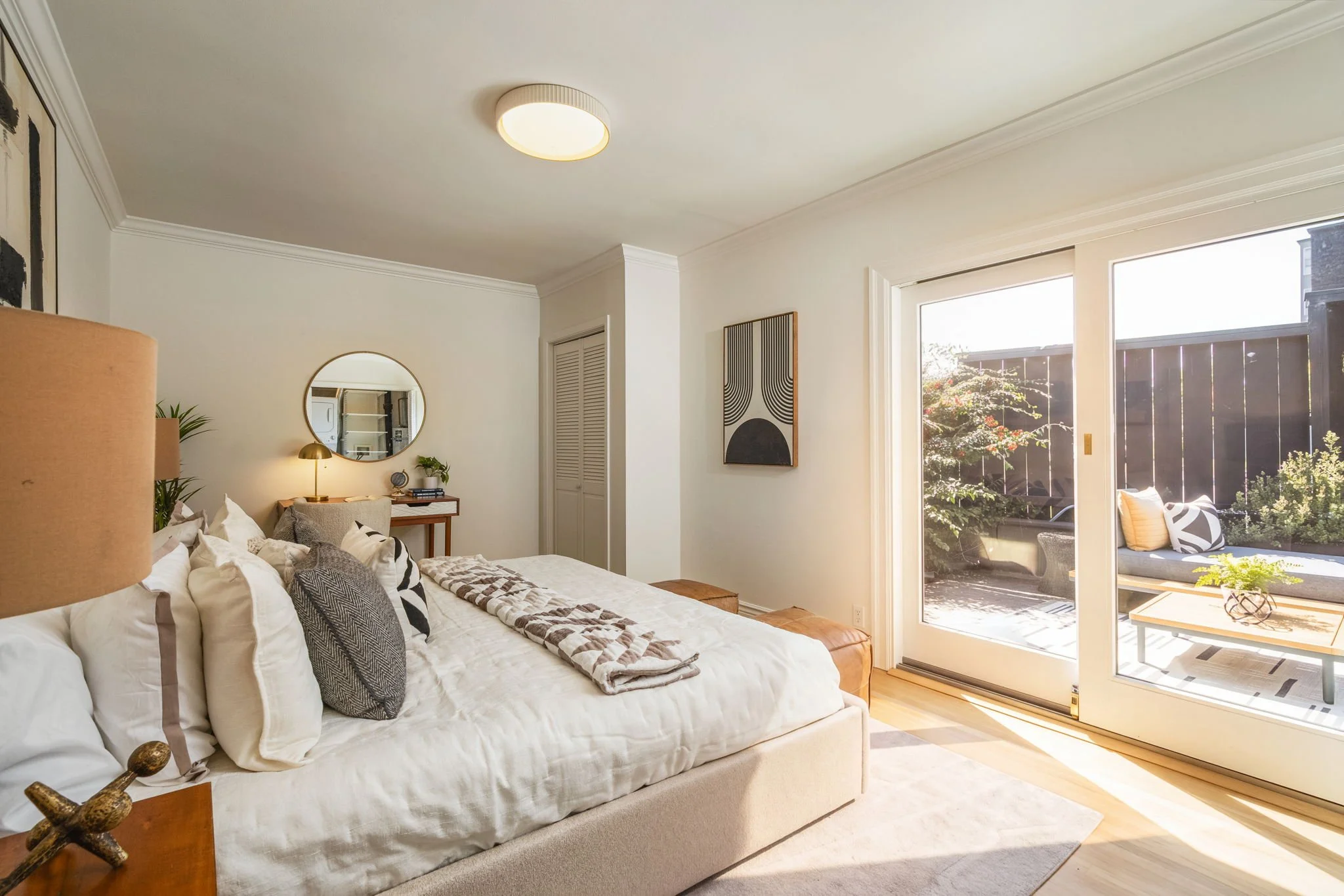 Bright bedroom with sliding glass door leading to outdoor patio, white walls, beige carpet, bed with white and patterned pillows, wall art, and a mirror.