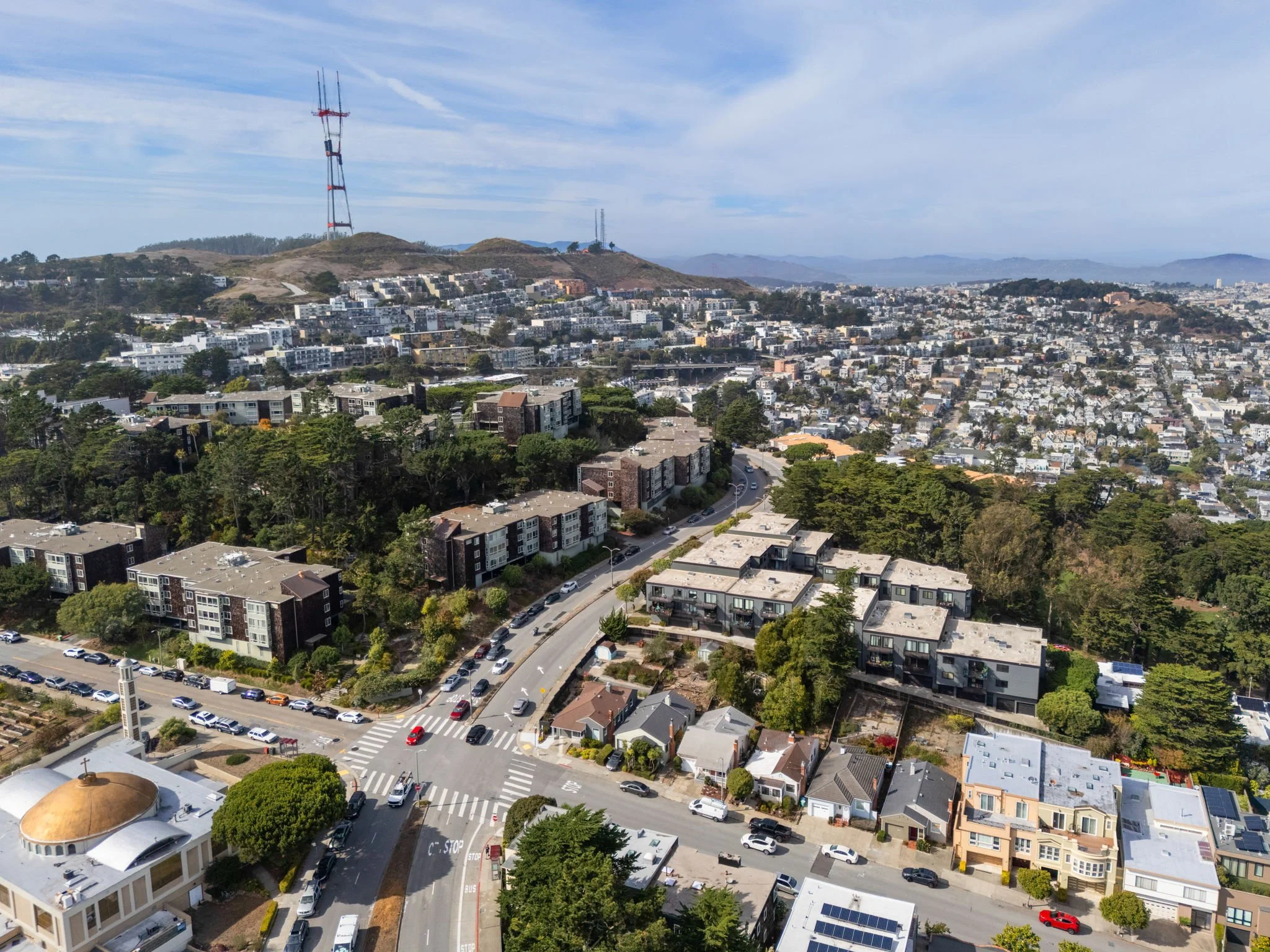 Aerial view of a residential neighborhood with houses and apartment buildings, a church, a small road intersection, and a hill with radio towers in the background under partly cloudy skies.