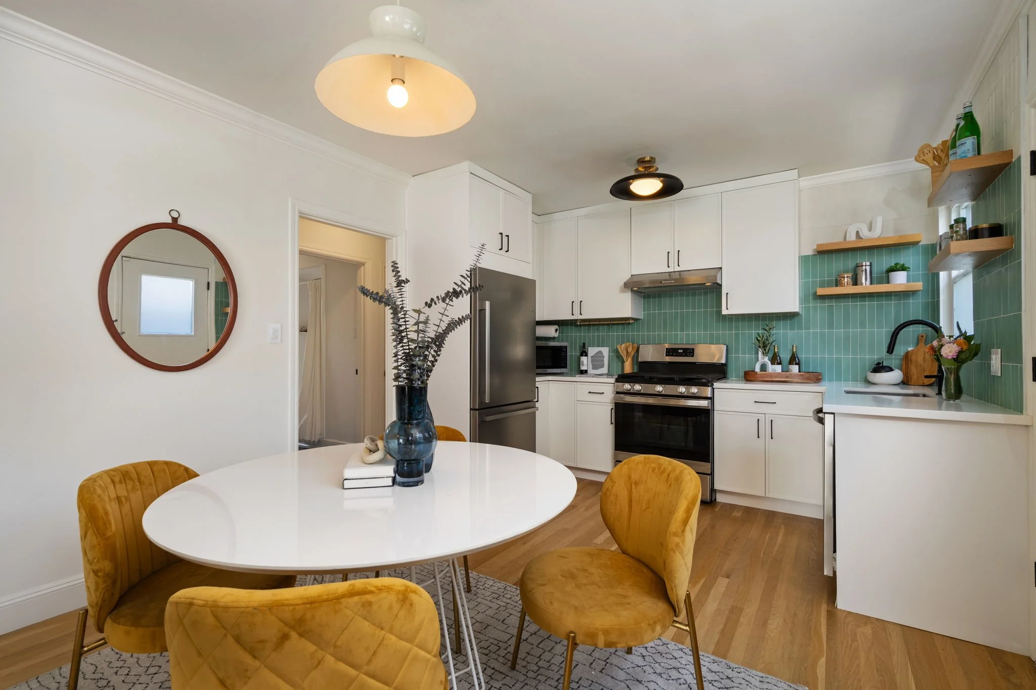 A modern kitchen and dining area with a white round table and four mustard yellow chairs. The kitchen has white cabinets, a stainless steel stove, and green backsplash tiles. Open wooden shelves hold bottles, plants, and decor. A large fish sculpture