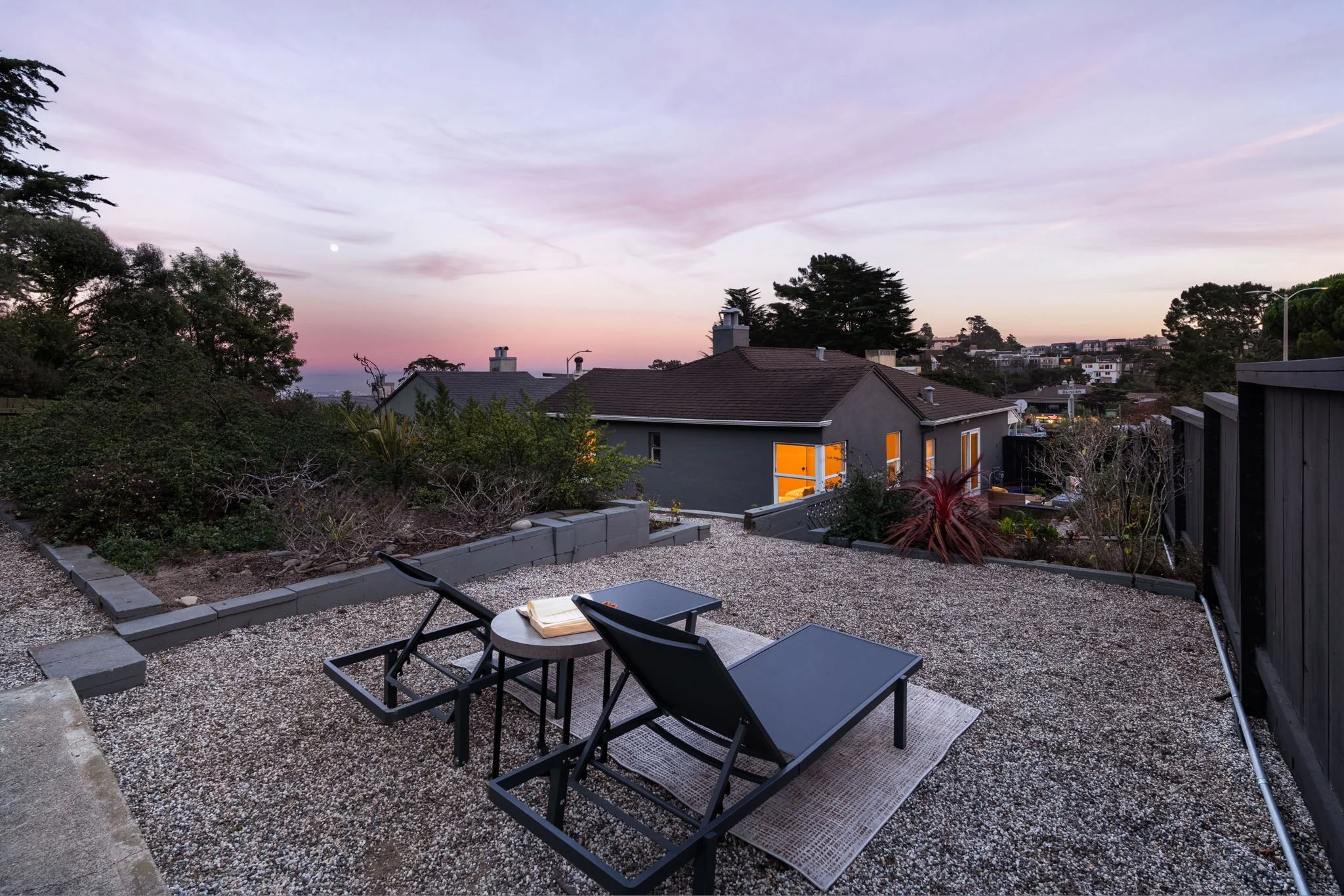 A backyard patio area with lounge chairs, a small table, and a book, overlooking neighboring houses and trees at dusk with a colorful sky.