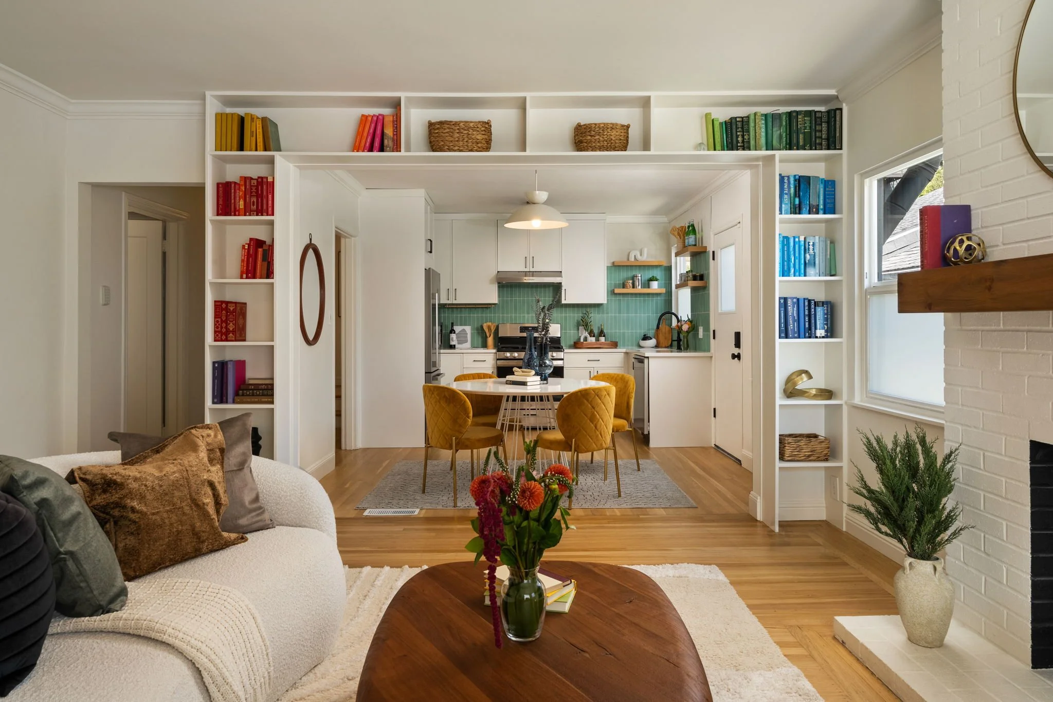 Open concept living room and kitchen with white walls, hardwood floors, and built-in bookshelves with colorful books. The living room has a white couch with pillows and a wooden coffee table with a vase of flowers. The kitchen features white cabinets, a green tiled backsplash, and a round dining table with yellow velvet chairs. There are decorative items, plants, and a window allowing natural light.