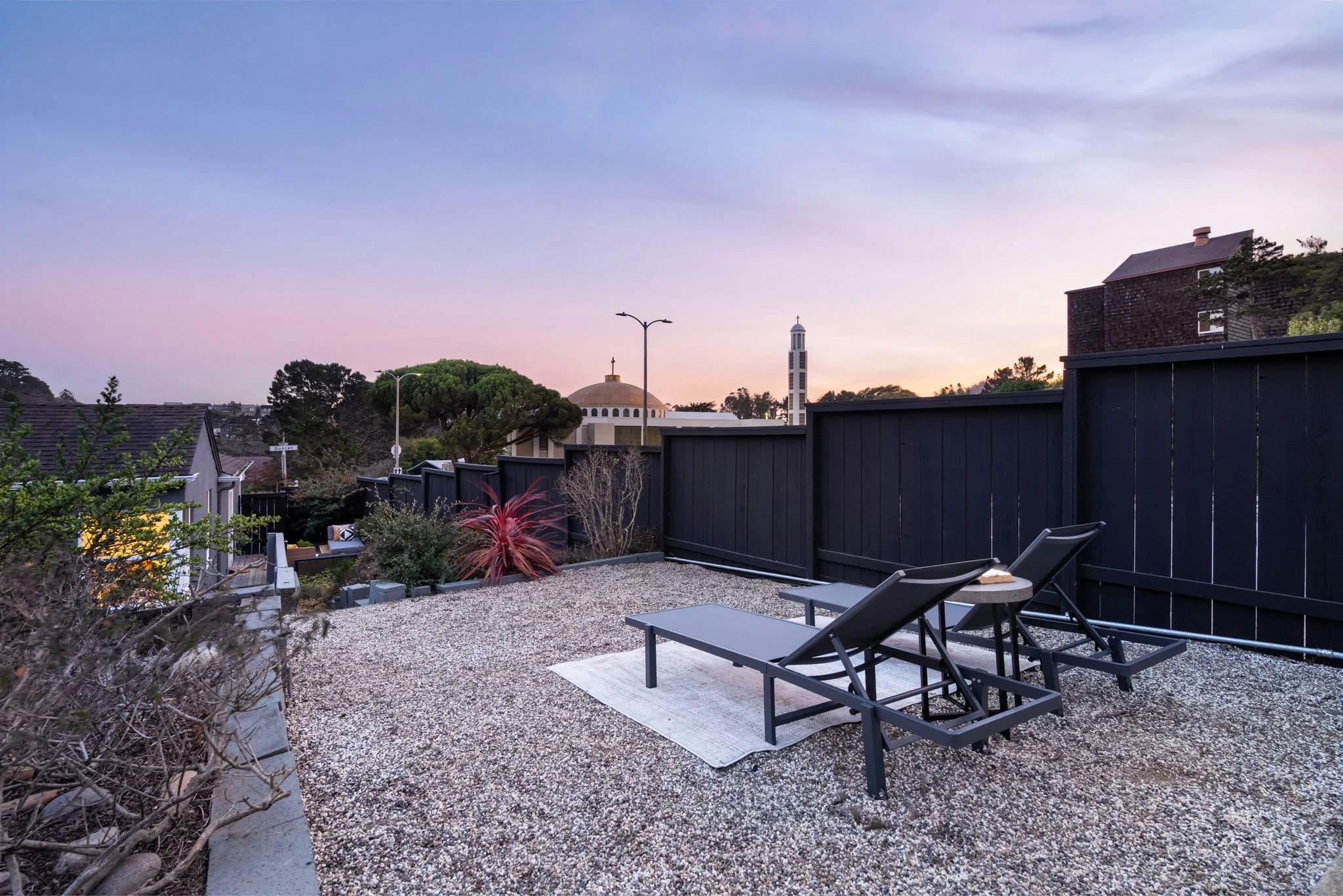 View of a backyard patio with two lounge chairs on a gravel surface, a small table, plants, and a black fence, overlooking a church with a dome and bell tower at sunset.