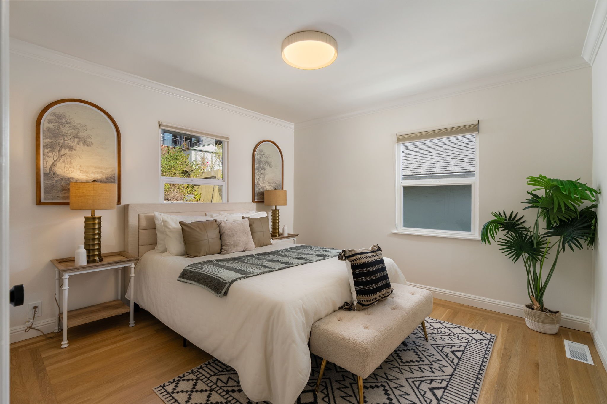 A bedroom with a white bed, two matching nightstands with lamps, a beige bench at the foot of the bed, and multiple windows with blinds. There is a large green plant in the corner.