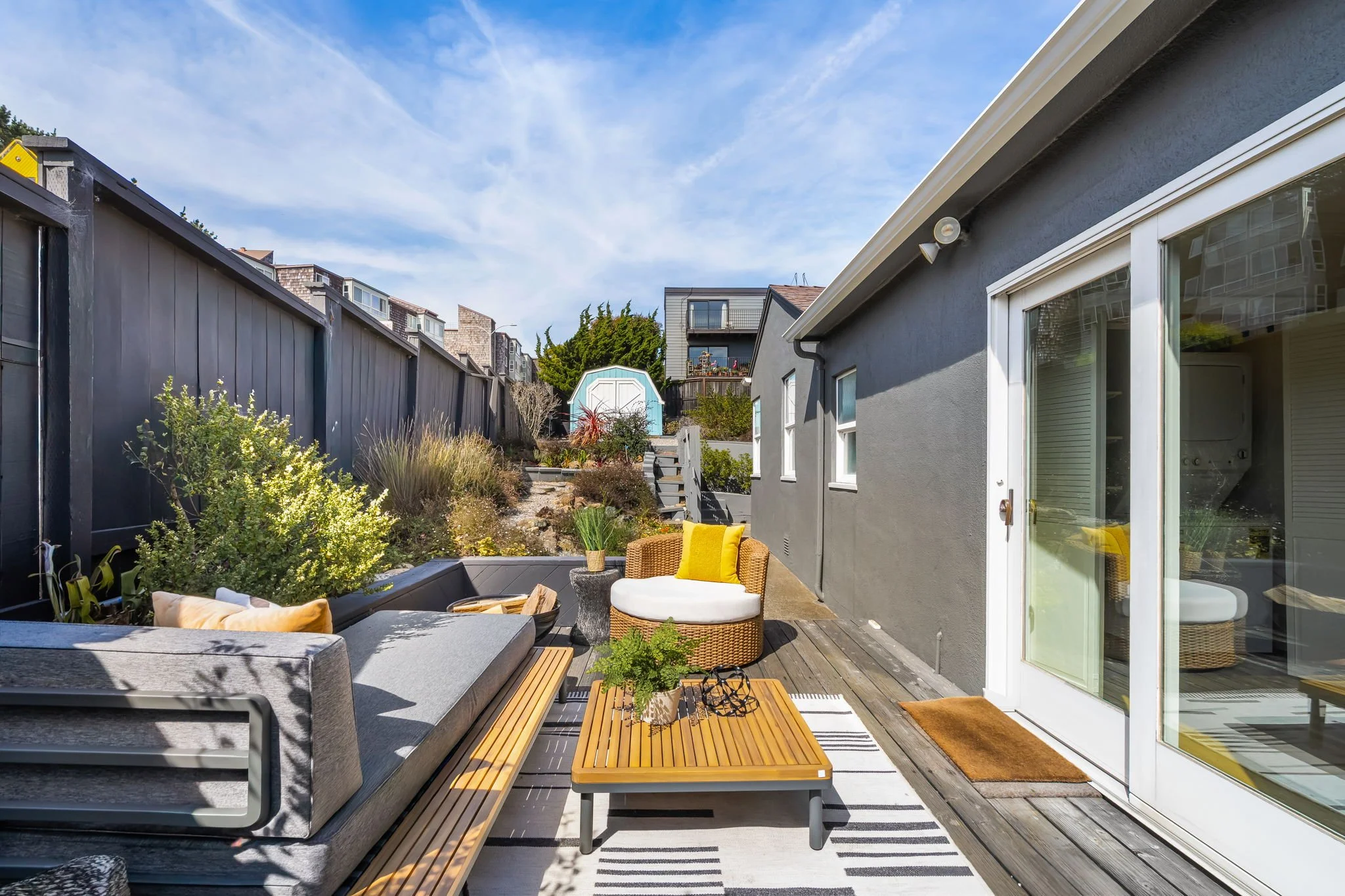 An outdoor patio area with seating, a wooden coffee table, and potted plants next to a gray house with large glass sliding doors under a clear blue sky.