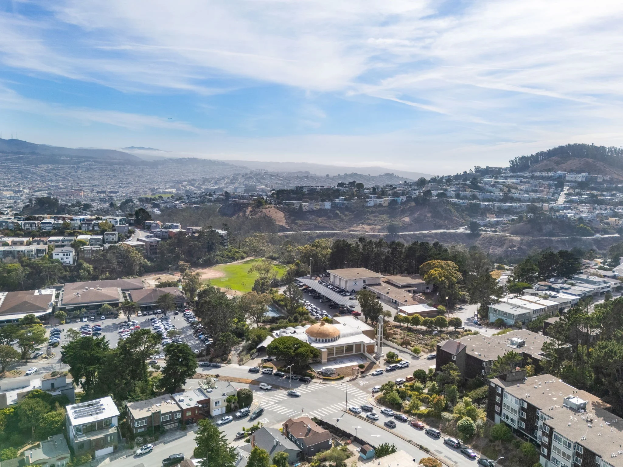 Aerial view of a city with residential buildings, a school or community center with a dome, a parking lot, and rolling hills in the background under a partly cloudy sky.