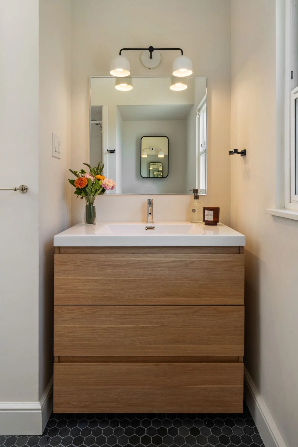 Modern bathroom vanity with a rectangular mirror, a wooden cabinet, a vase of colorful flowers, and bathroom essentials under a light fixture.