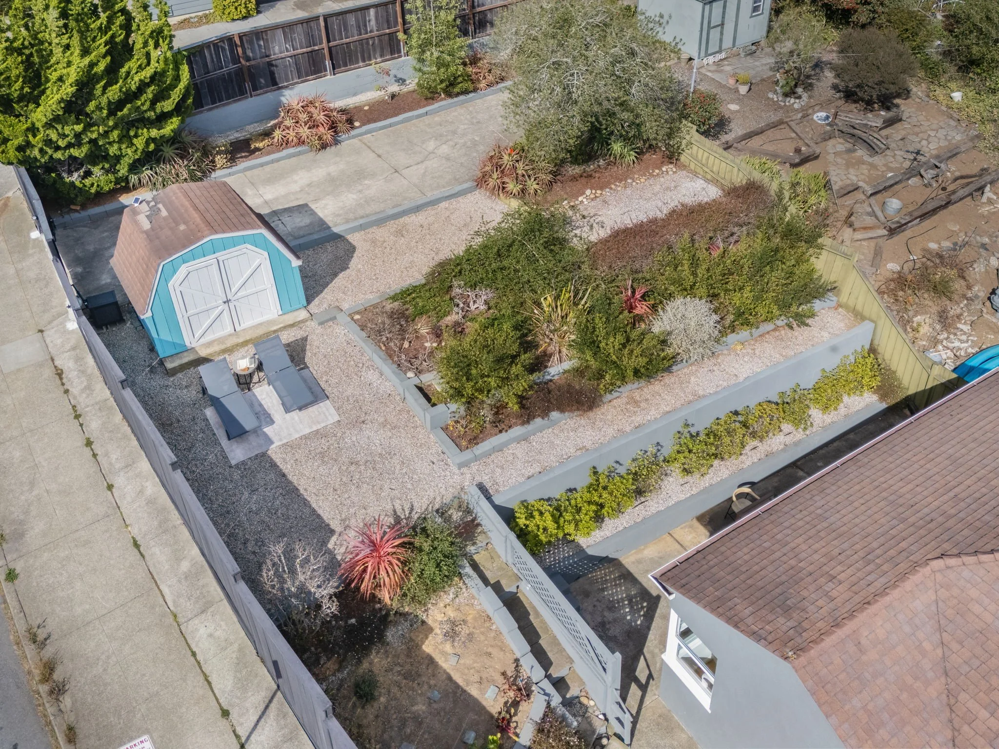 A bird's-eye view of a residential backyard with trees, shrubs, a small shed, a patio, gravel, and a retaining wall with a climbing plant, bordered by a driveway and neighboring houses.