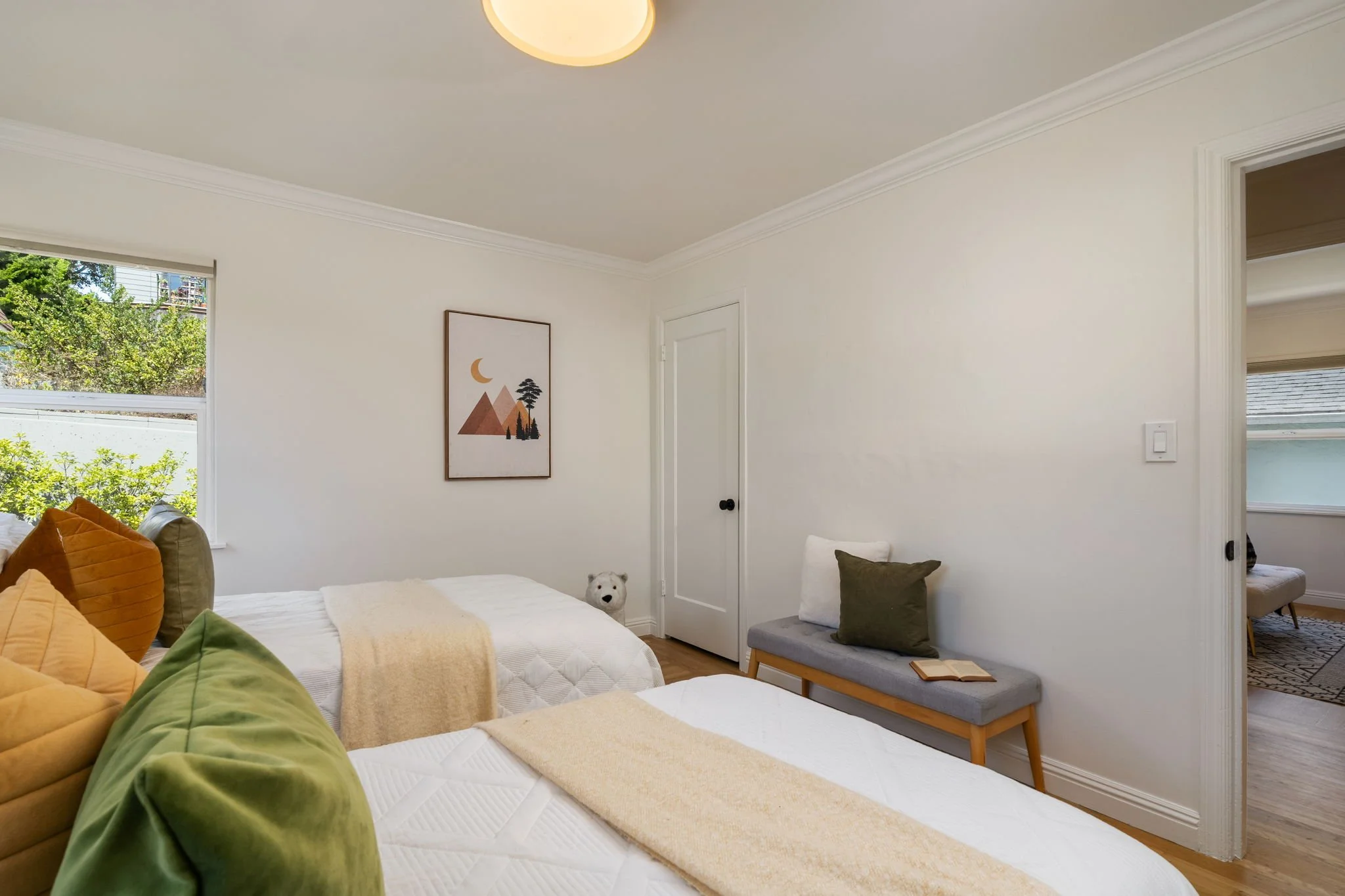 A bedroom with white walls and crown molding. There are multiple beds with white bedding and colorful pillows in shades of green, orange, and brown. A window shows green trees outside. A framed minimalist mountain and moon print hangs on the wall. Th