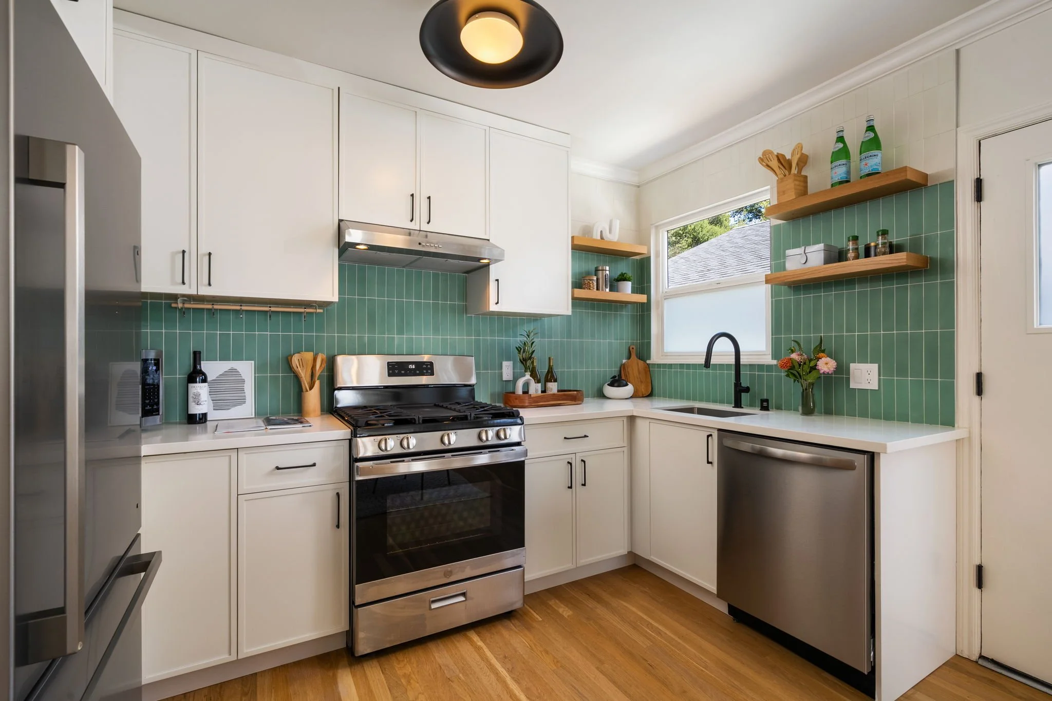 A modern kitchen with white cabinets, green tile backsplash, stainless steel appliances, wooden open shelves, a black kitchen faucet, and a window.