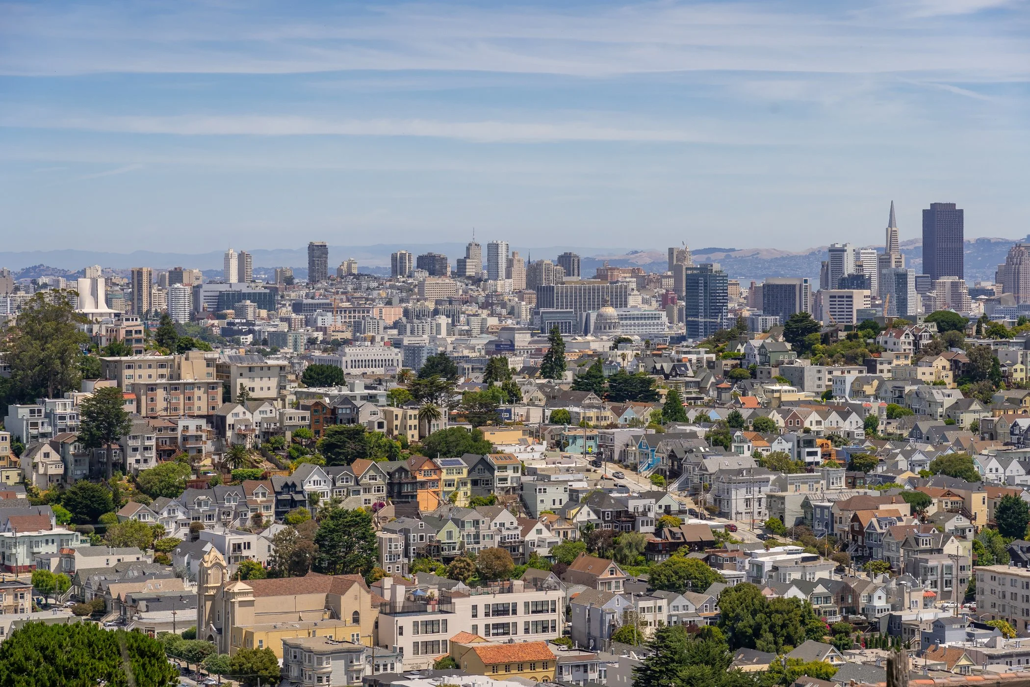 A panoramic cityscape of San Francisco with numerous high-rise buildings, the Transamerica Pyramid, and rolling hills in the background, under a partly cloudy sky.