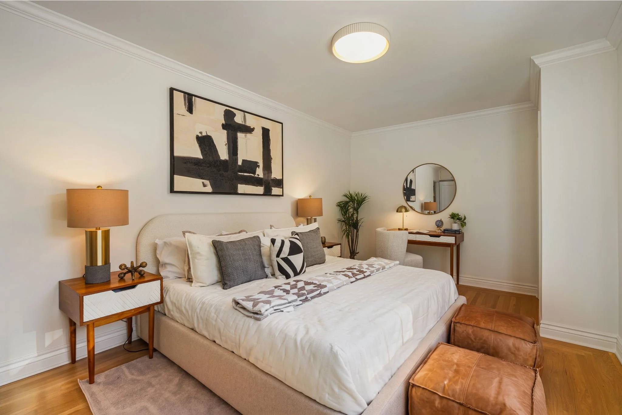 A bedroom with a beige upholstered bed, white bedding, and decorative pillows. A wooden nightstand with a modern lamp and small decorative objects. Artwork hanging above the bed, a mirror, a small desk with lamps, plants, and a leather ottoman at the