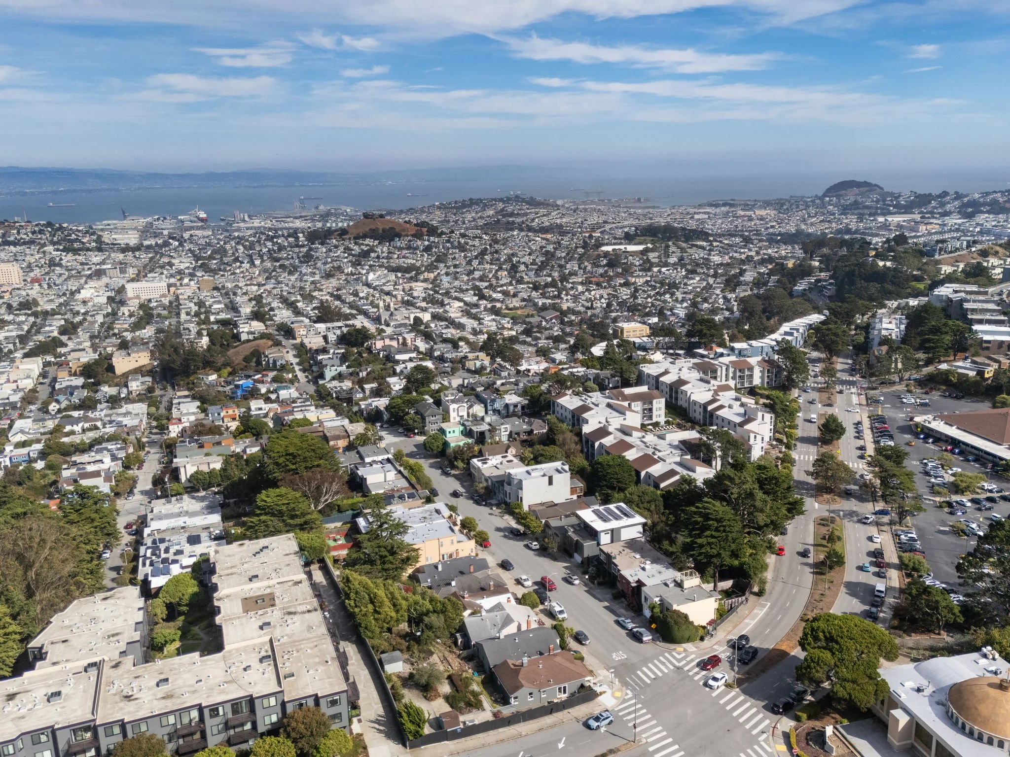 An aerial view of a densely populated urban area with numerous residential houses, trees, and roads, overlooking a body of water with ships and a hill in the background.
