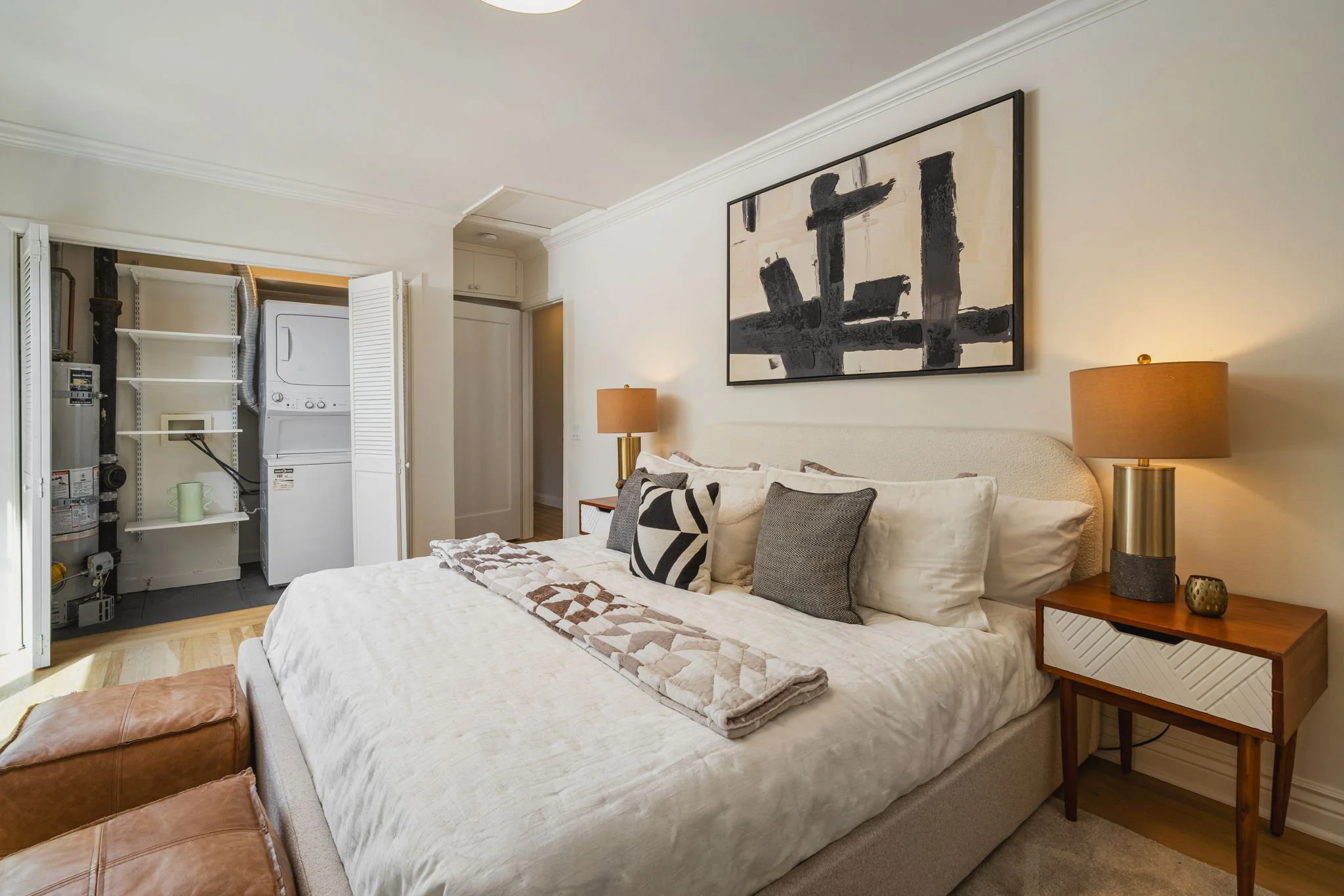 A bedroom with a large bed, nightstand with lamp, abstract black and white artwork on the wall, and an open closet with laundry appliances.