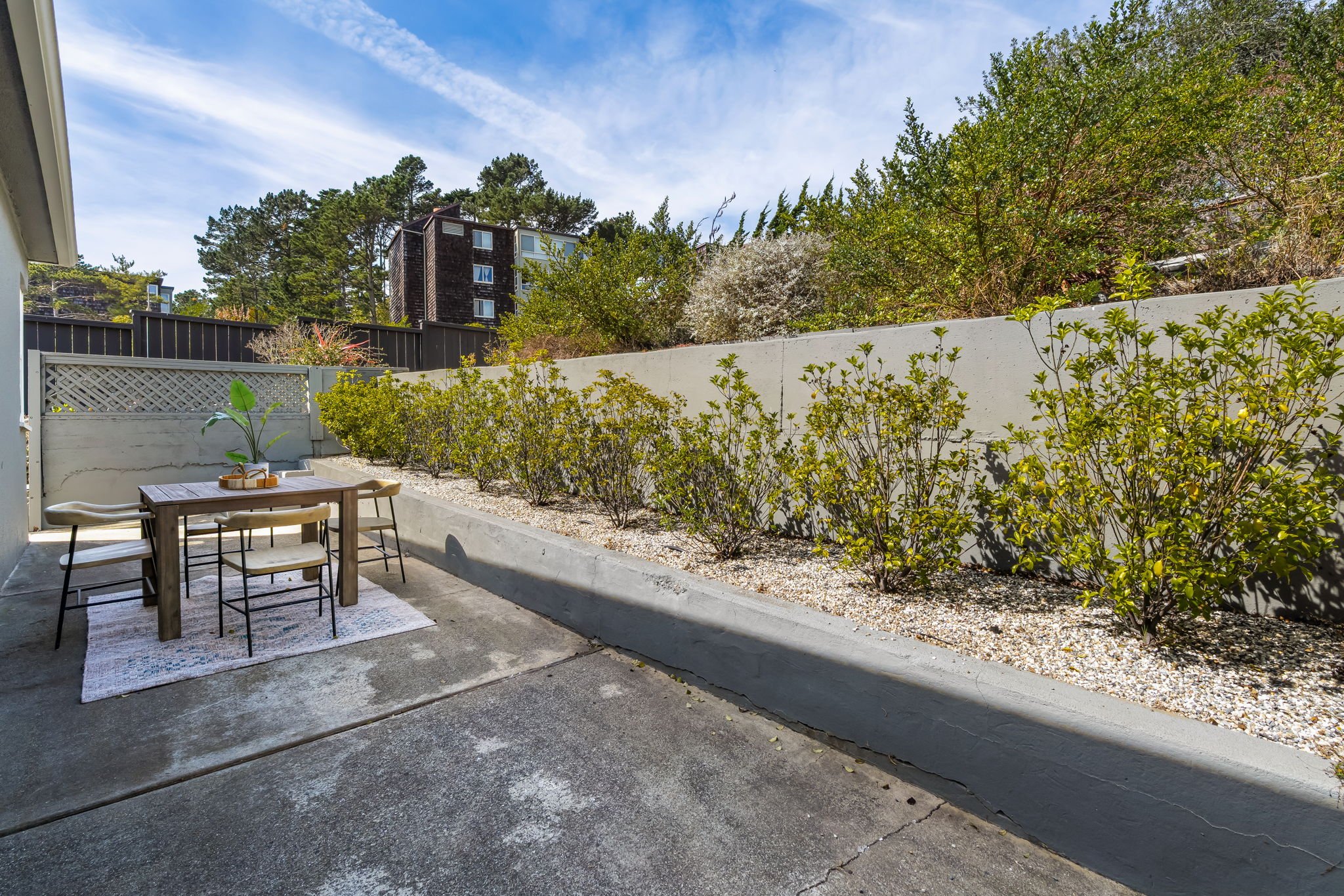 A backyard patio with a wooden dining table, four chairs, and a rug, surrounded by a low wall with bushes. There are trees and a multi-story building in the background under a partly cloudy sky.