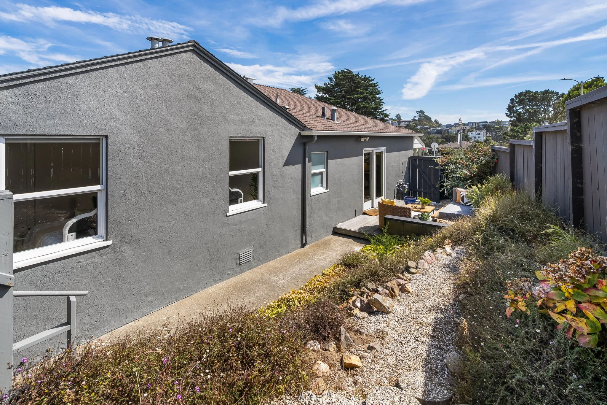 Exterior backyard scene with gray house wall, windows, patio furniture, garden area with plants and rocks, and neighboring houses on a hill under a partly cloudy sky.