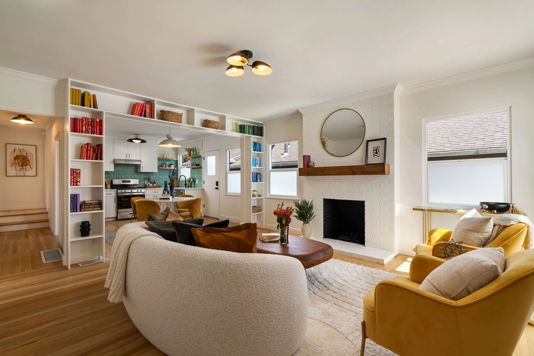 Living room with white brick fireplace, yellow chairs, beige sofa, wooden coffee table, built-in bookshelves with colorful books, and windows with blinds.
